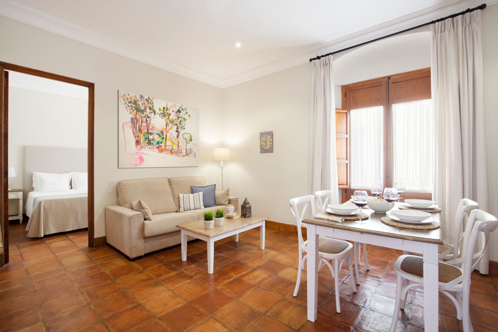 Bright living-dining room with beige sofa, white table, hardwood floor, and large windows with curtains