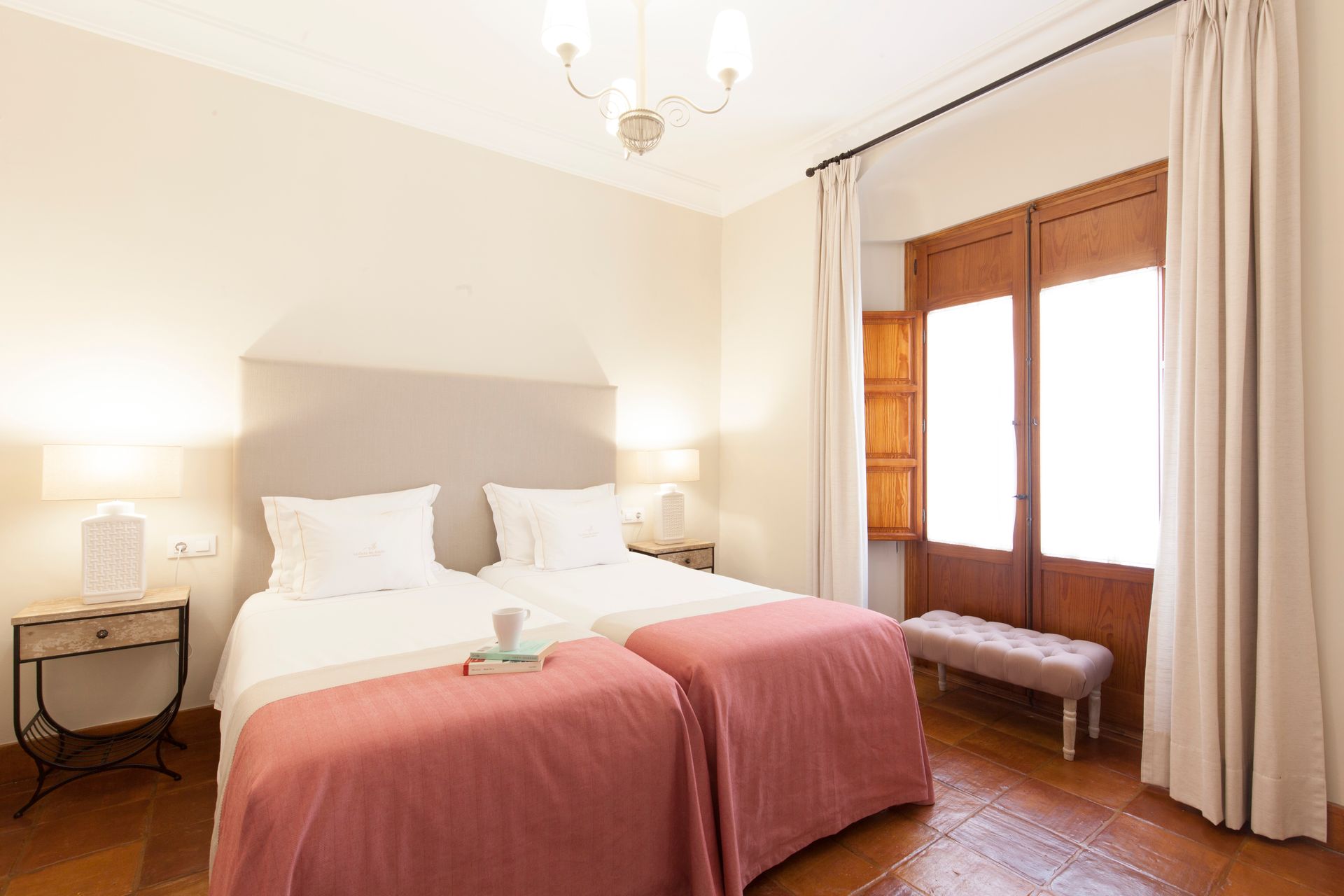 Bright bedroom with two white beds, pink blankets, bedside lamps, and a window seat by the window