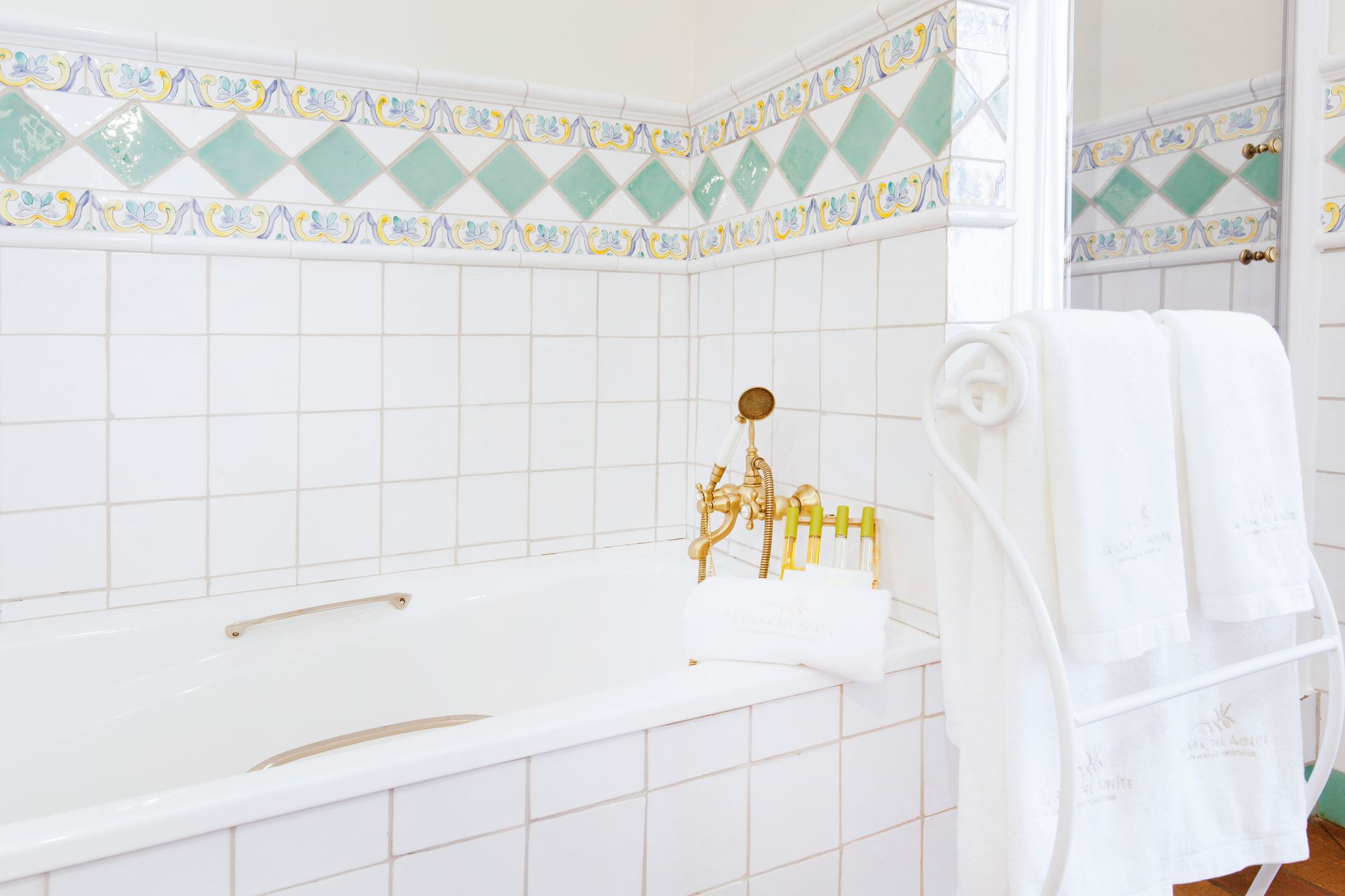 Bright bathroom with white tiles, a white tub, gold faucet, and towels on a rack