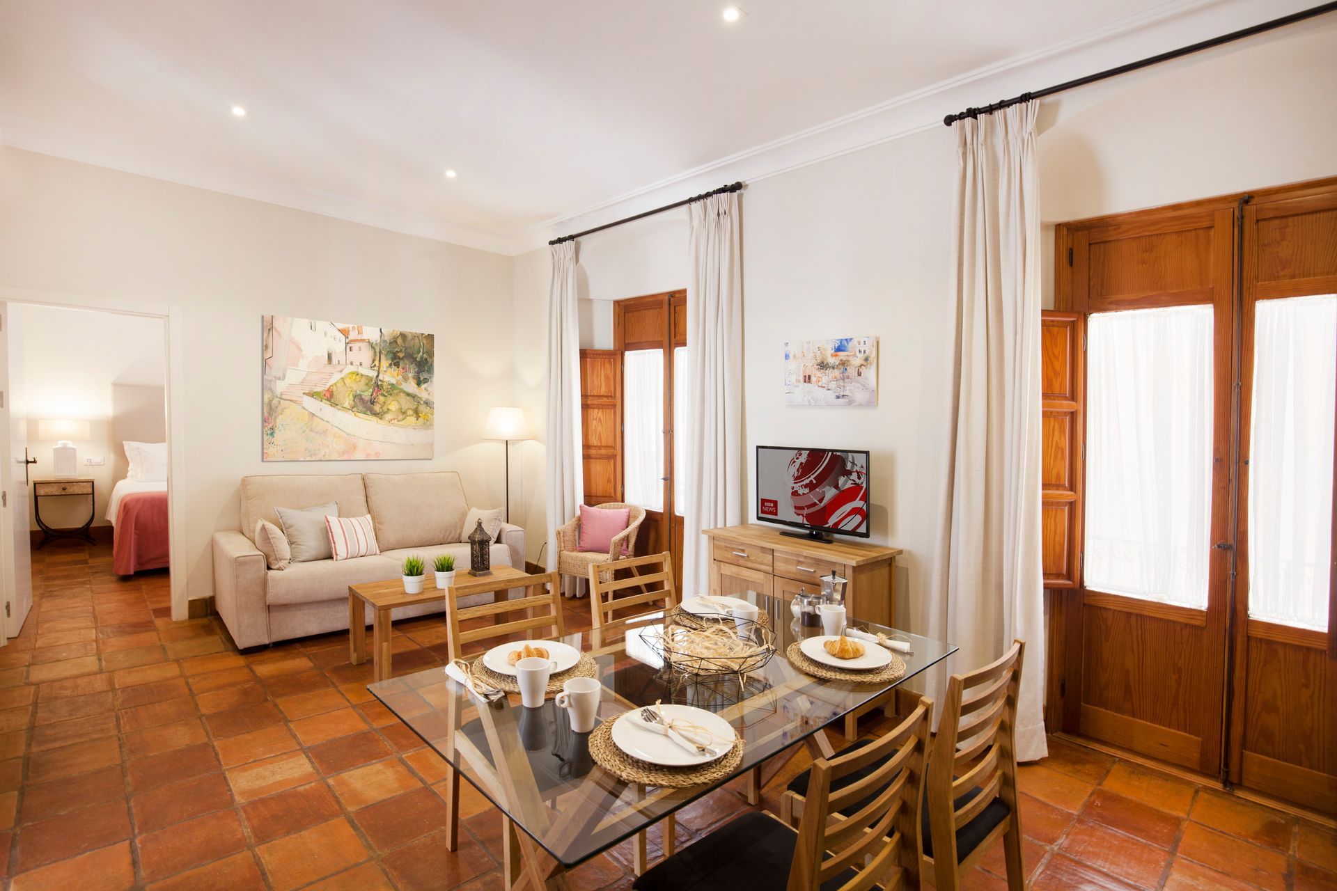 Bright dining room with wooden table, beige sofa, terracotta tile floor, and tall windows with white curtains