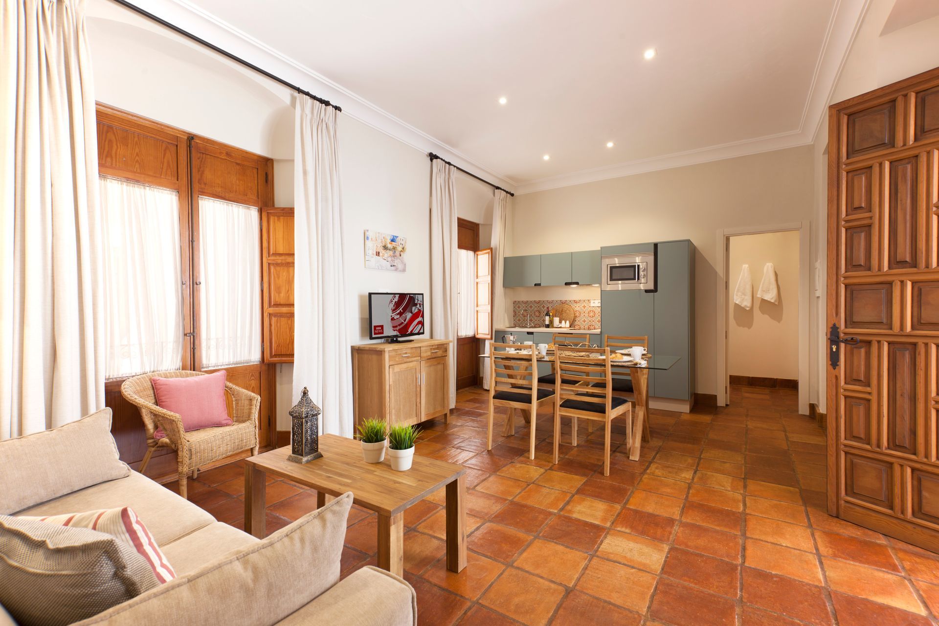 Bright living room with terracotta tile, cream curtains, and a dining table leading to a kitchen area