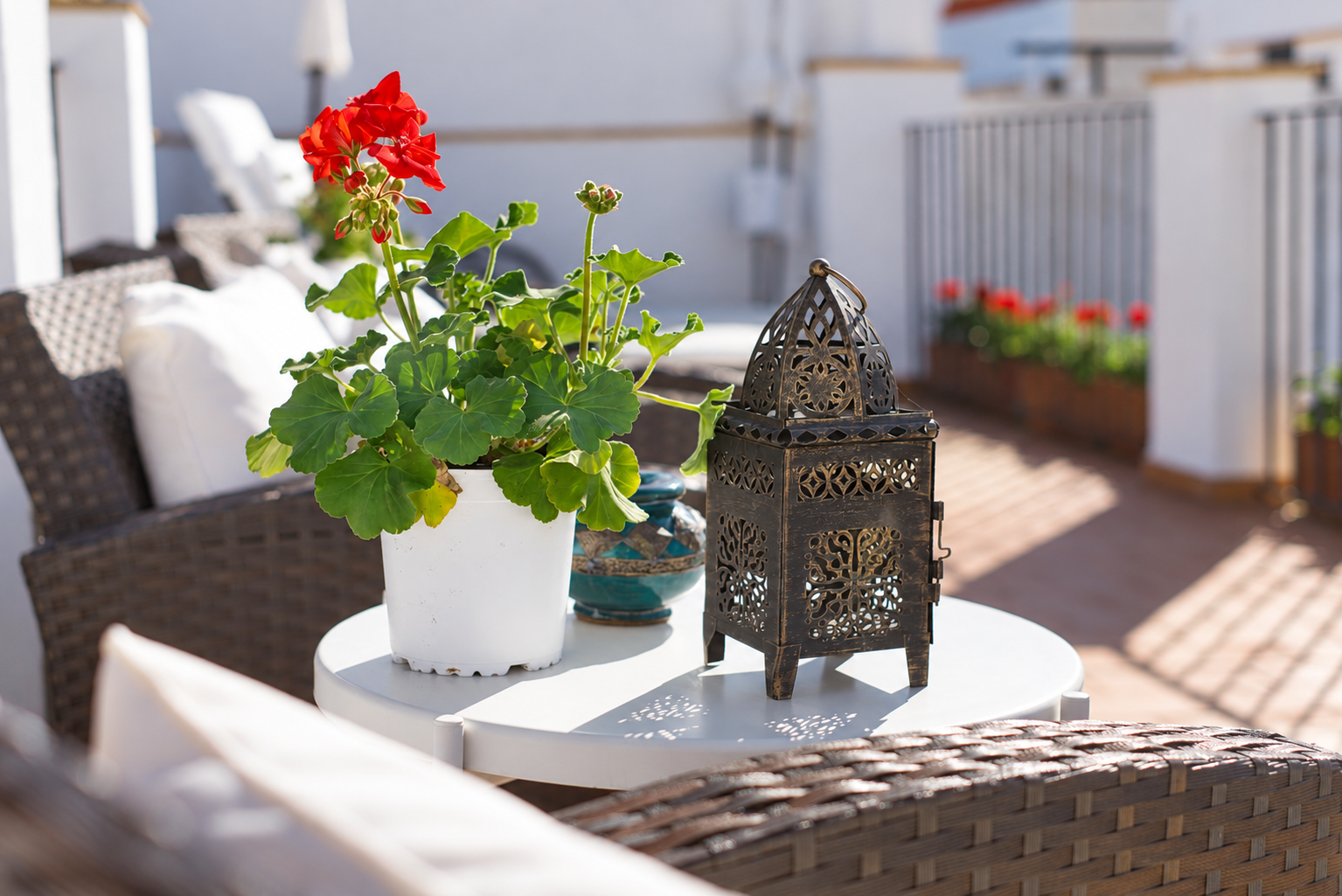White patio table with potted red flowers and a decorative metal lantern in a sunny outdoor seating area