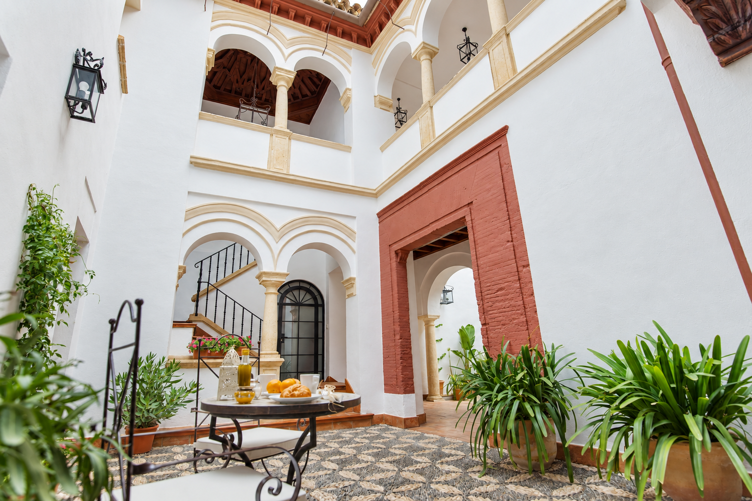 Bright courtyard with white arches, red trim, patterned tile floor, and potted plants around a small table.