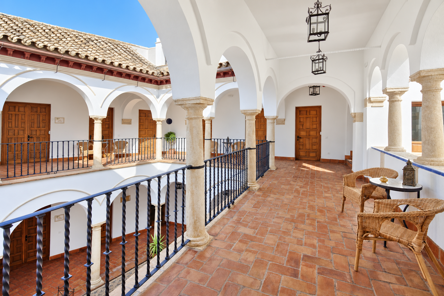 Sunlit Spanish-style courtyard with white arches, terracotta tiles, and wooden doors on an upper balcony.