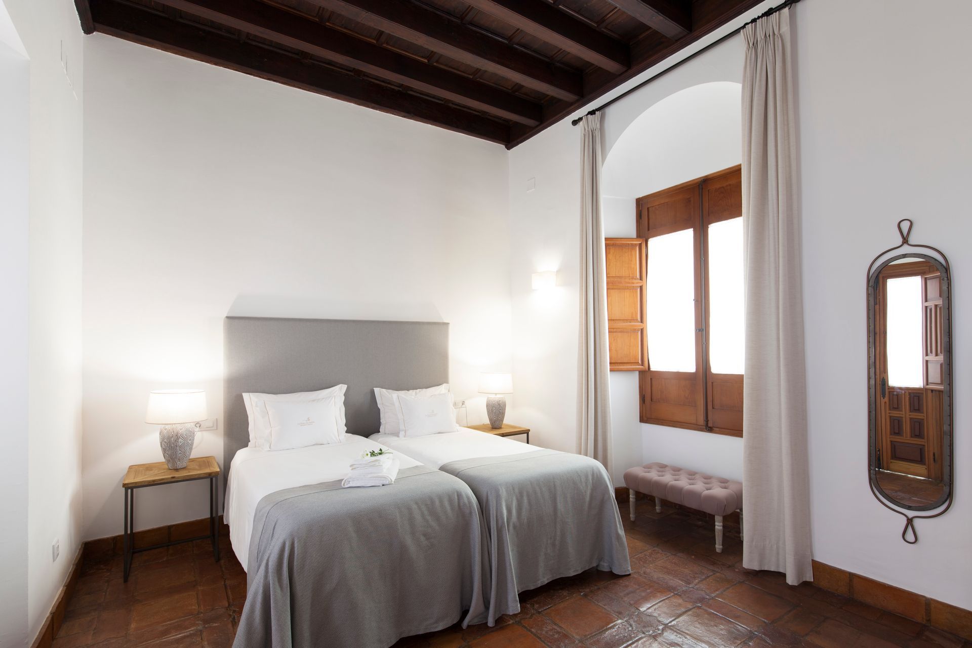 A cozy bedroom with a white bed, gray throw, wooden ceiling beams, bedside lamps, and a tall window with curtains.