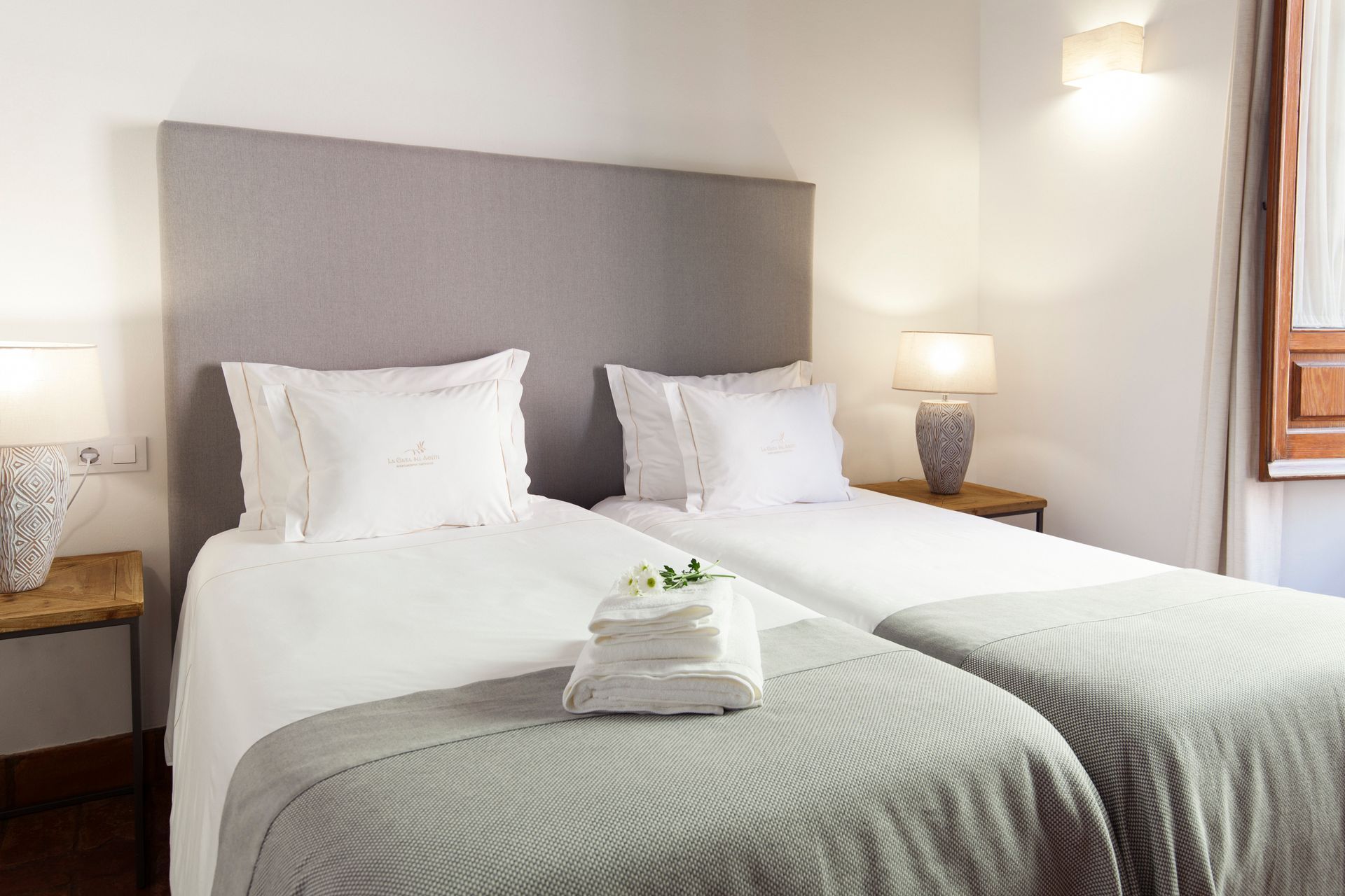 Bright bedroom with two white twin beds, gray headboards, bedside lamps, and a window.