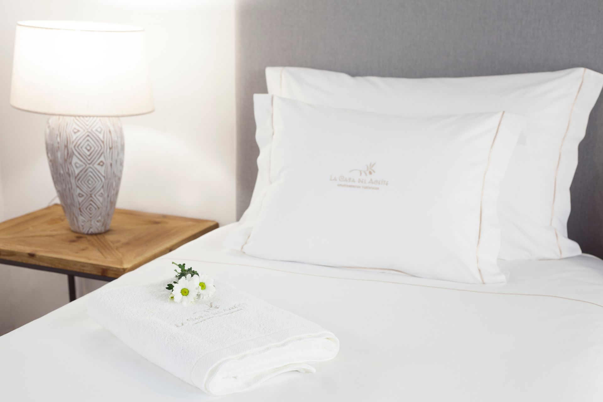 White bed with folded towels and flowers beside a wooden table and lamp in a bright bedroom