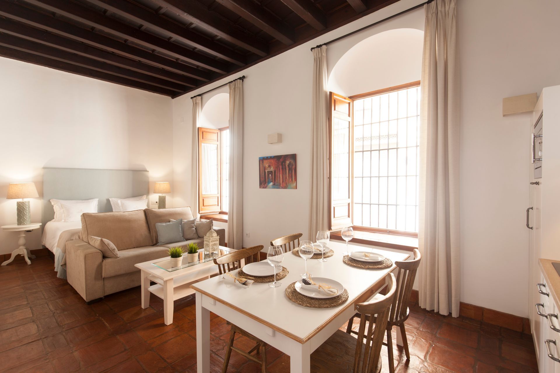 Bright dining and living room with white table, beige sofa, tall windows, and wooden ceiling beams