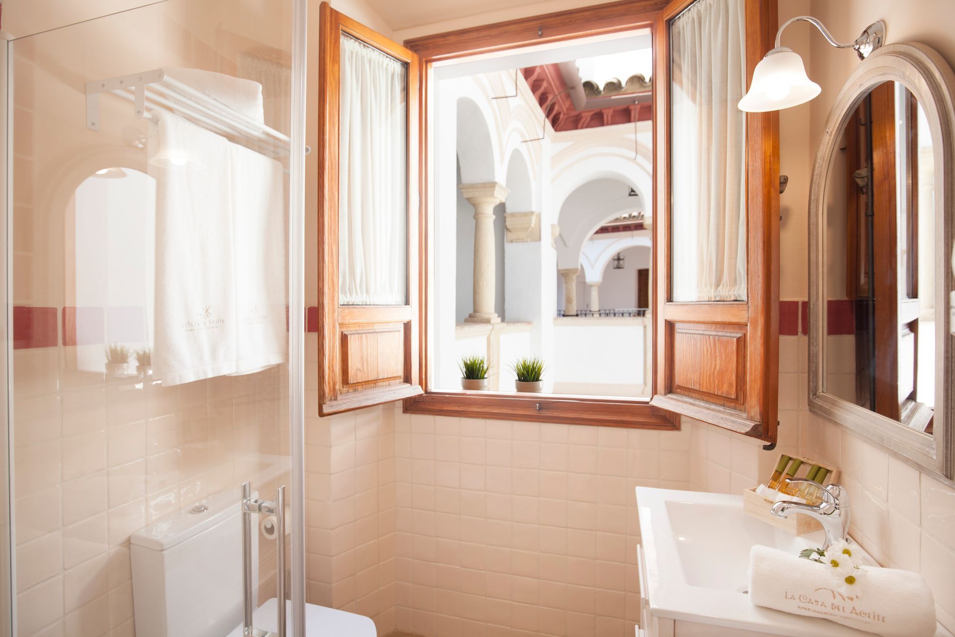 Bright bathroom with open wooden windows, white sink, mirror, and view of an arched courtyard outside