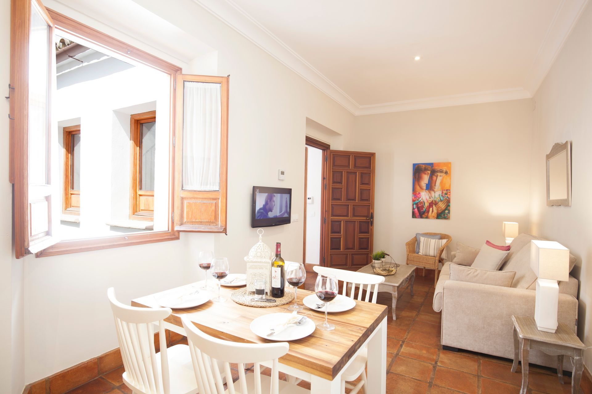 Bright dining-living room with white chairs, set table, sofa, wall art, and open window shutters