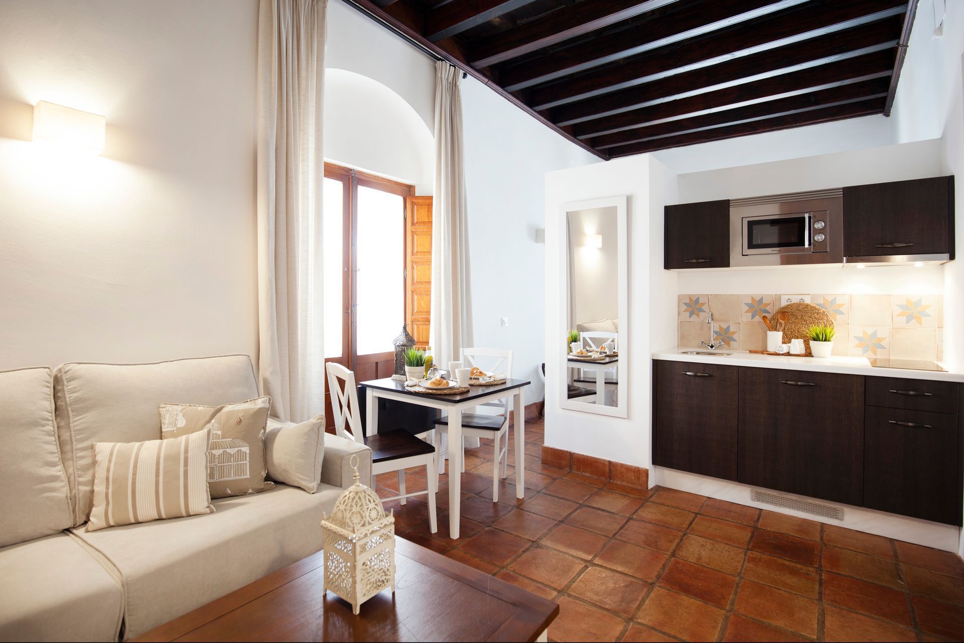 Cozy open-plan living room and kitchen with beige sofa, dark wood cabinets, and terracotta tile floor.