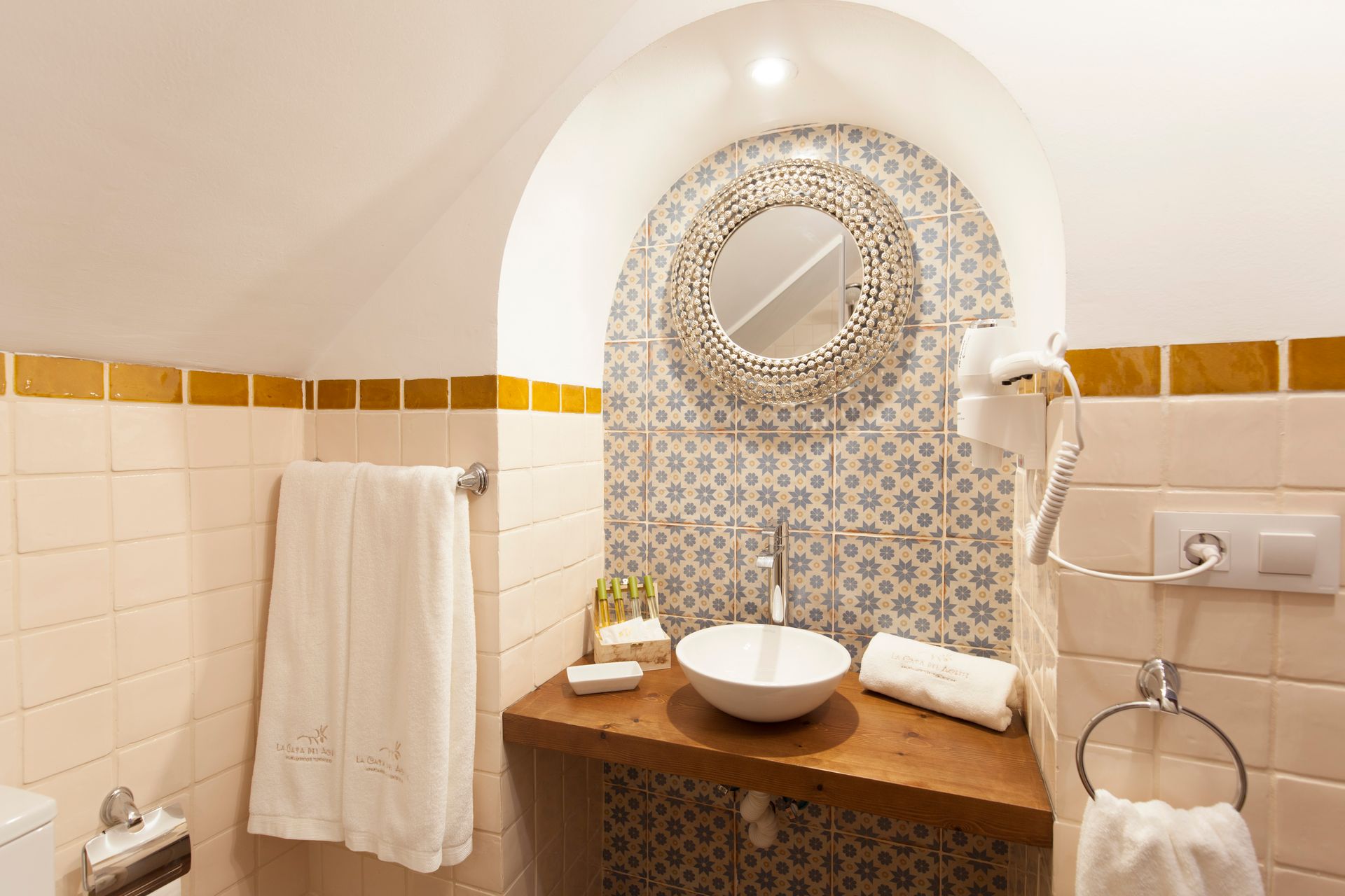 Small tiled bathroom with arched vanity, round mirror, vessel sink, towel, and wall-mounted hair dryer.