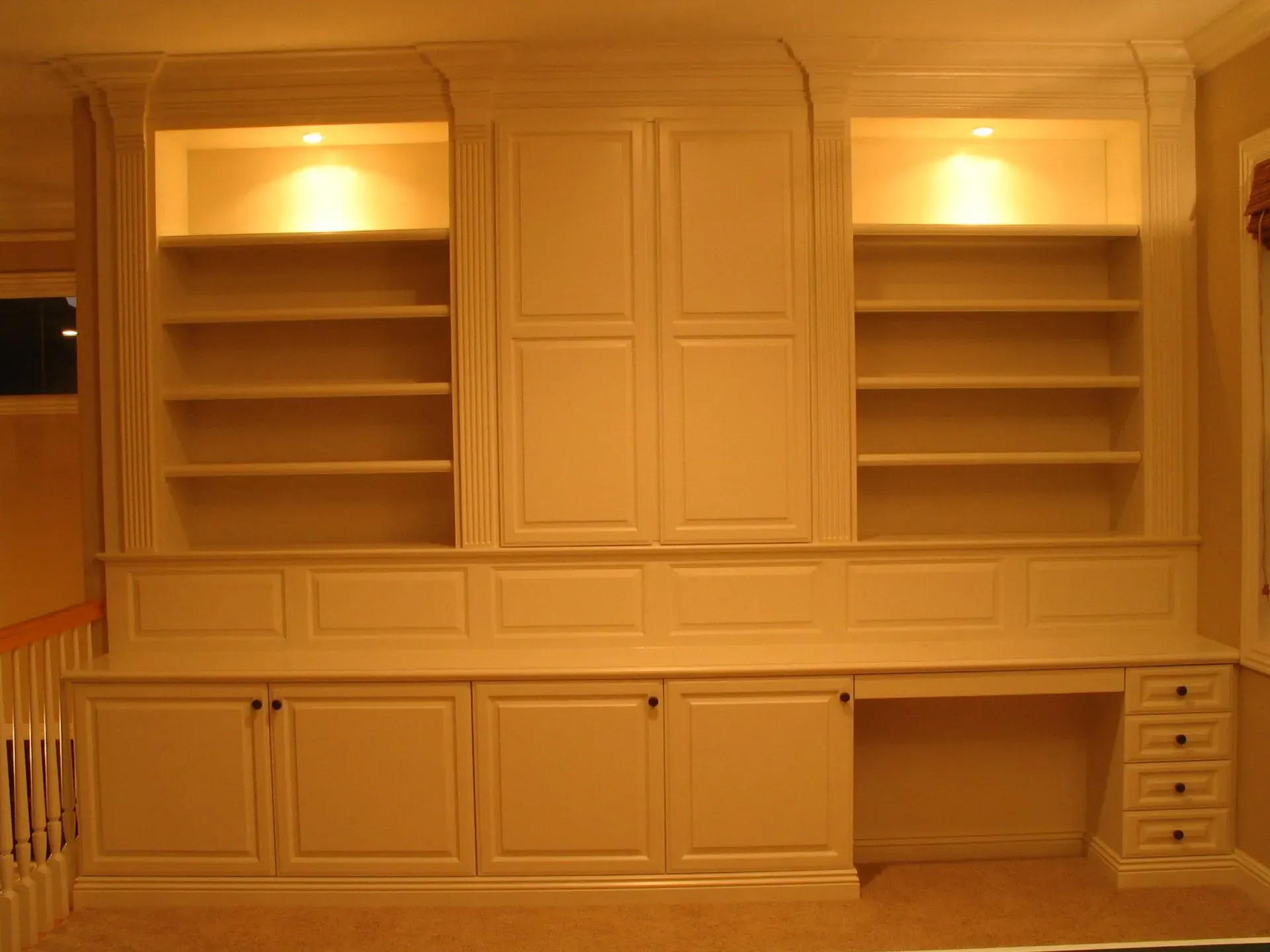 Built-in white wood wall unit with shelves, cabinets, and lit display niches in a warm room