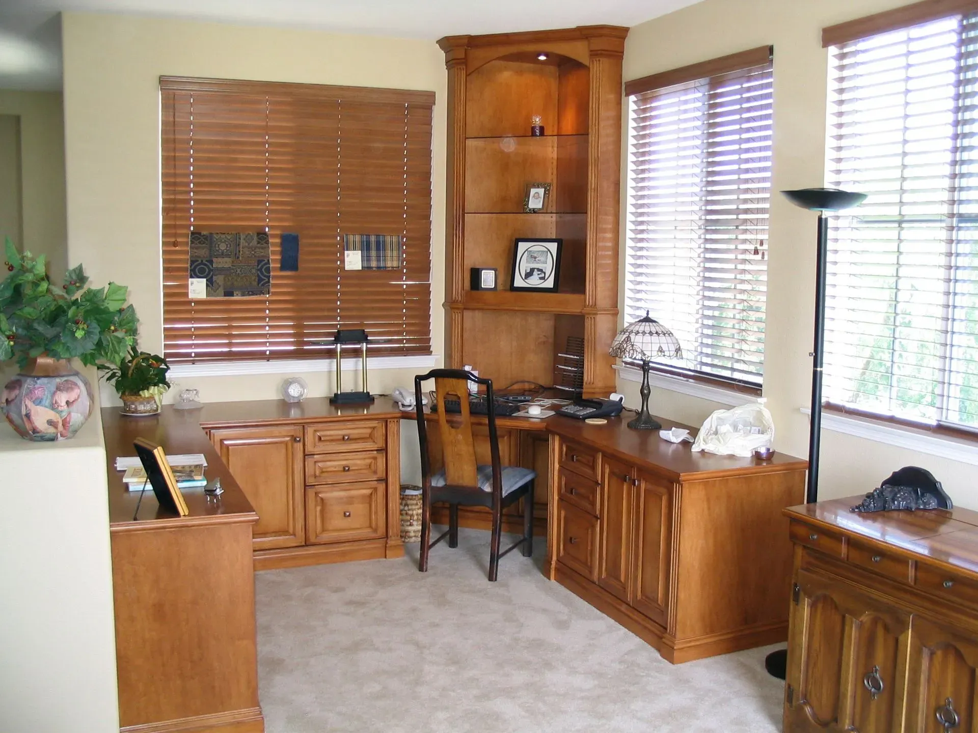 Home office with wooden desk, corner shelves, blinds, and a chair by the window.
