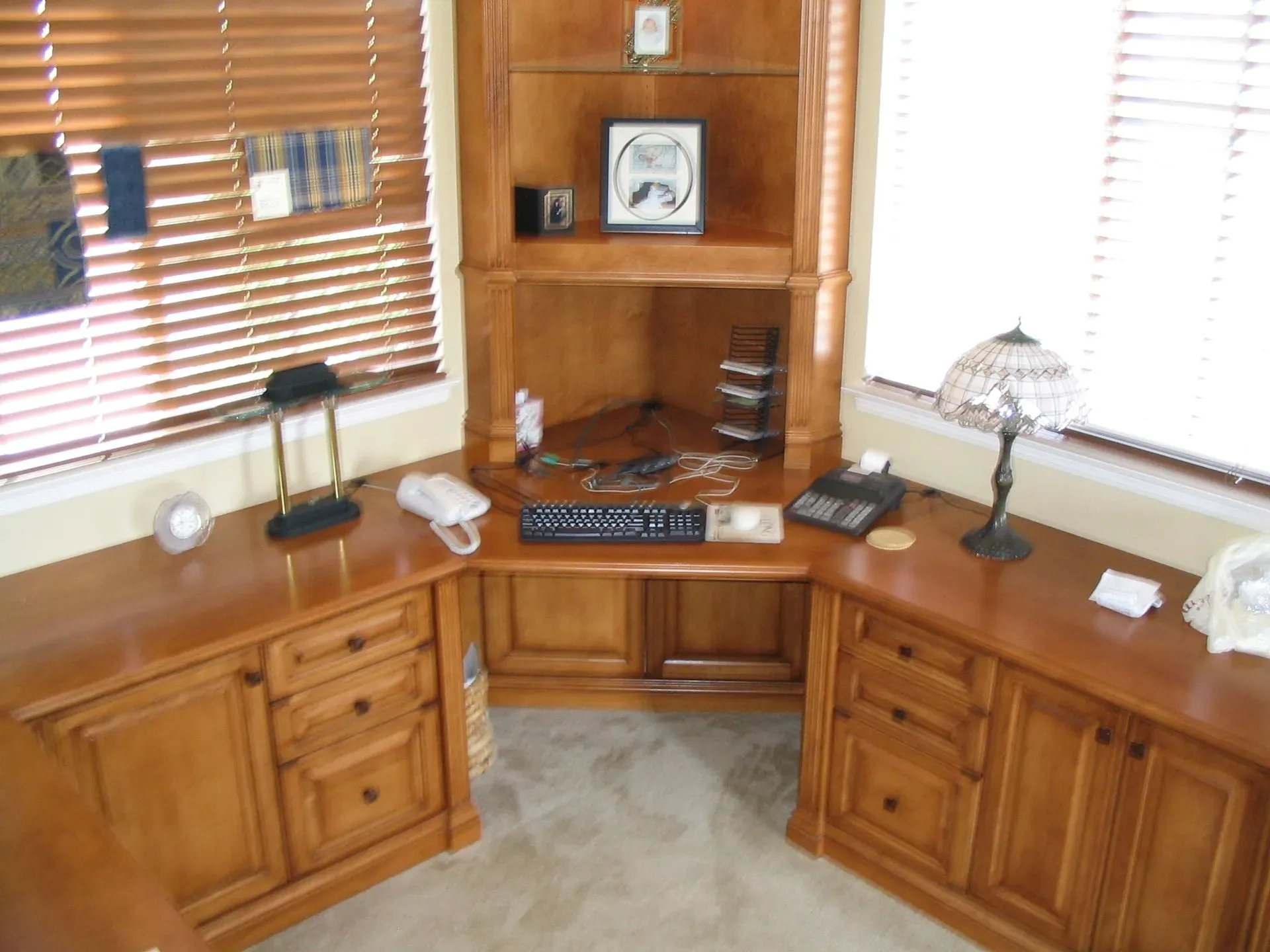Wood-paneled home office corner with built-in desk, monitor, phone, lamp, and blinds over the windows.