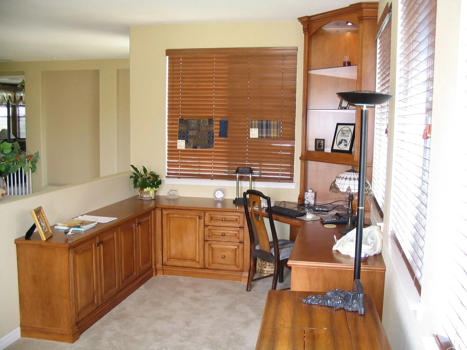 Home office corner with wood desk, cabinets, and shelves beside a window with blinds.