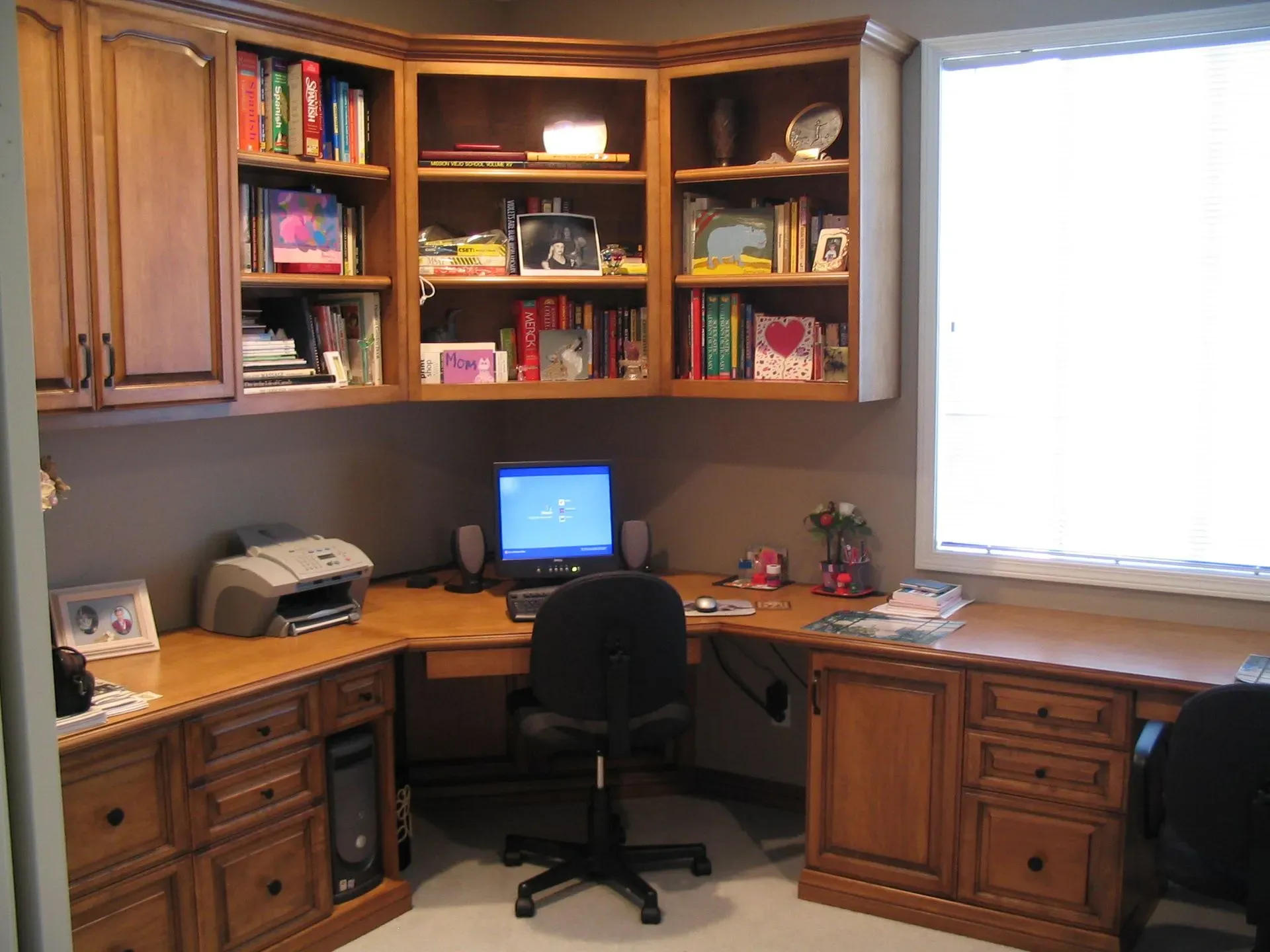 Corner home office with wooden desk, shelves, a computer, and a chair by a bright window.