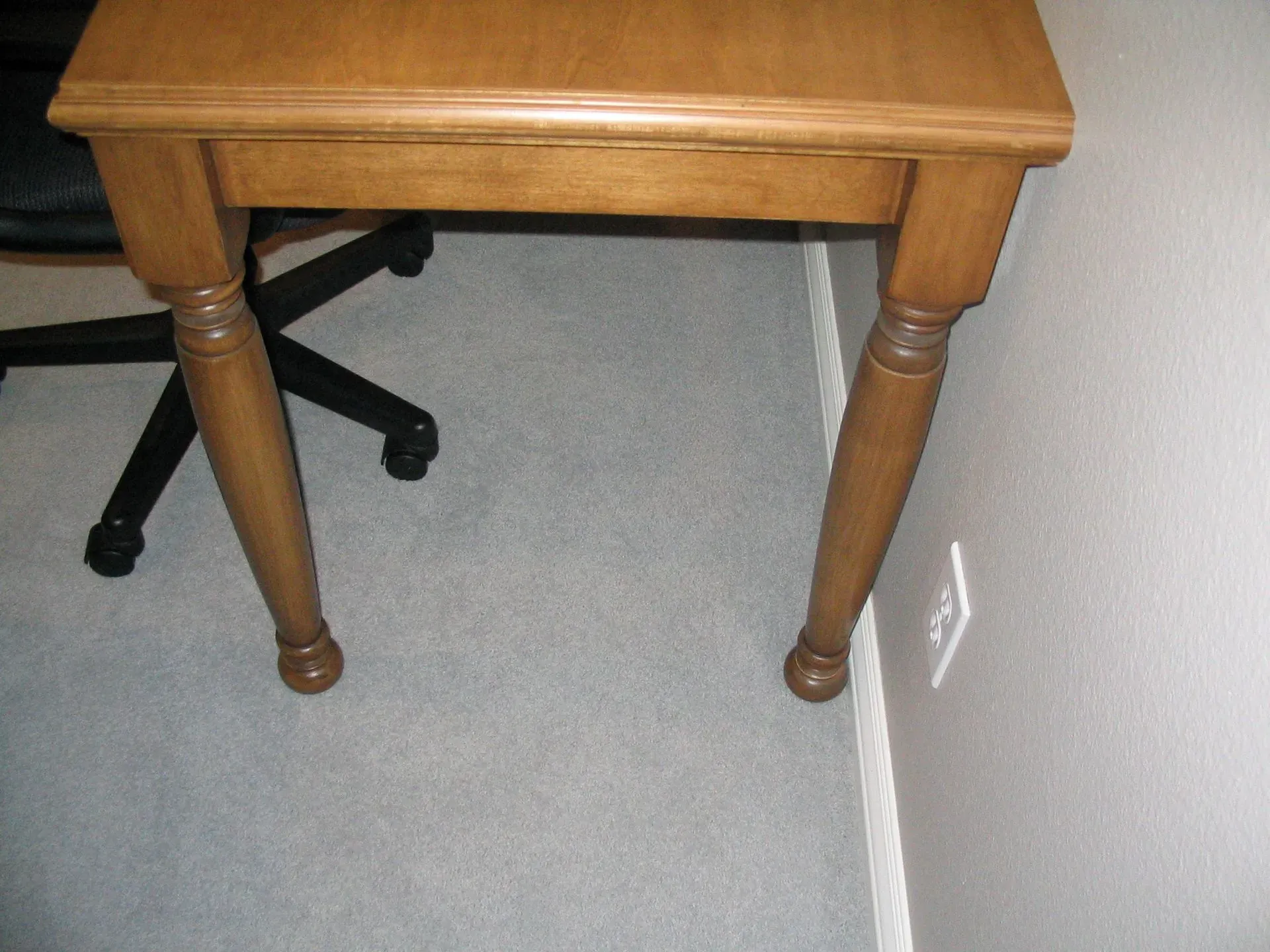 Small wooden table beside a black office chair on a gray carpet near a wall outlet.