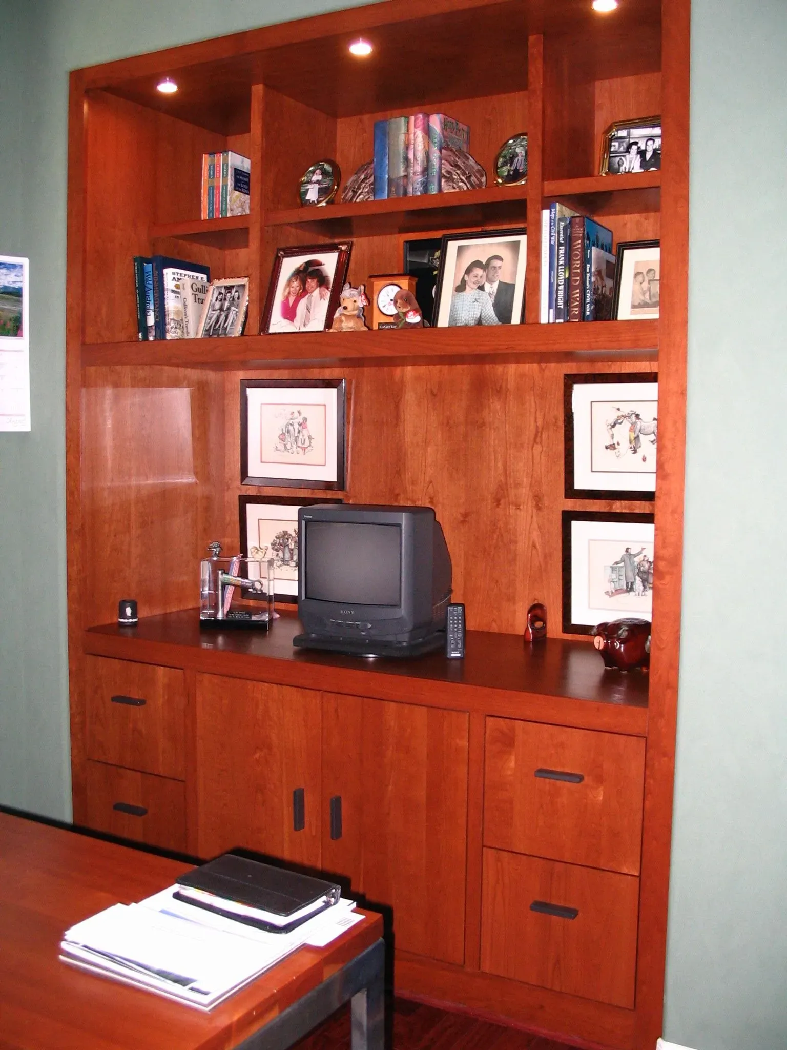 Wooden built-in office cabinet with shelves, books, framed photos, and a small TV on a desk