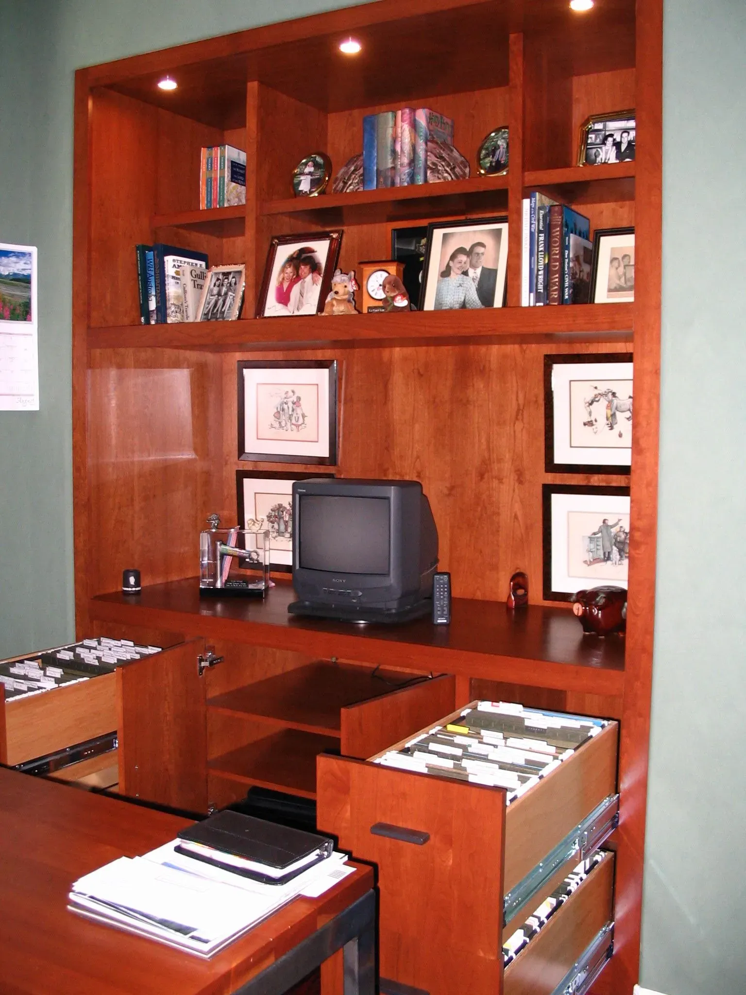 Wooden home office wall unit with shelves, framed photos, books, and a small TV above a desk.