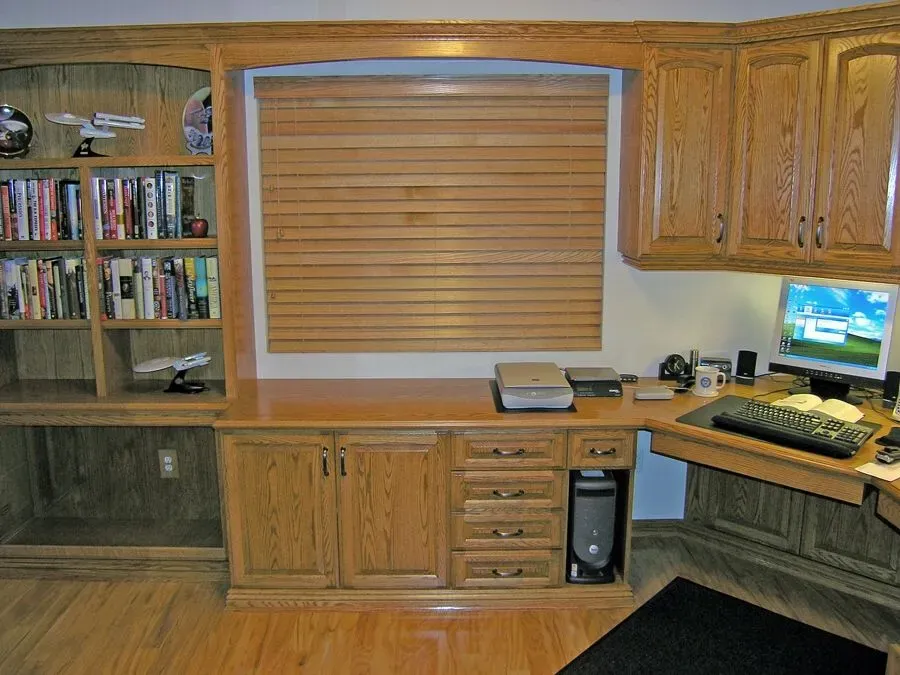 Wooden home office with built-in desk, bookshelves, cabinets, computer, and a window with blinds.
