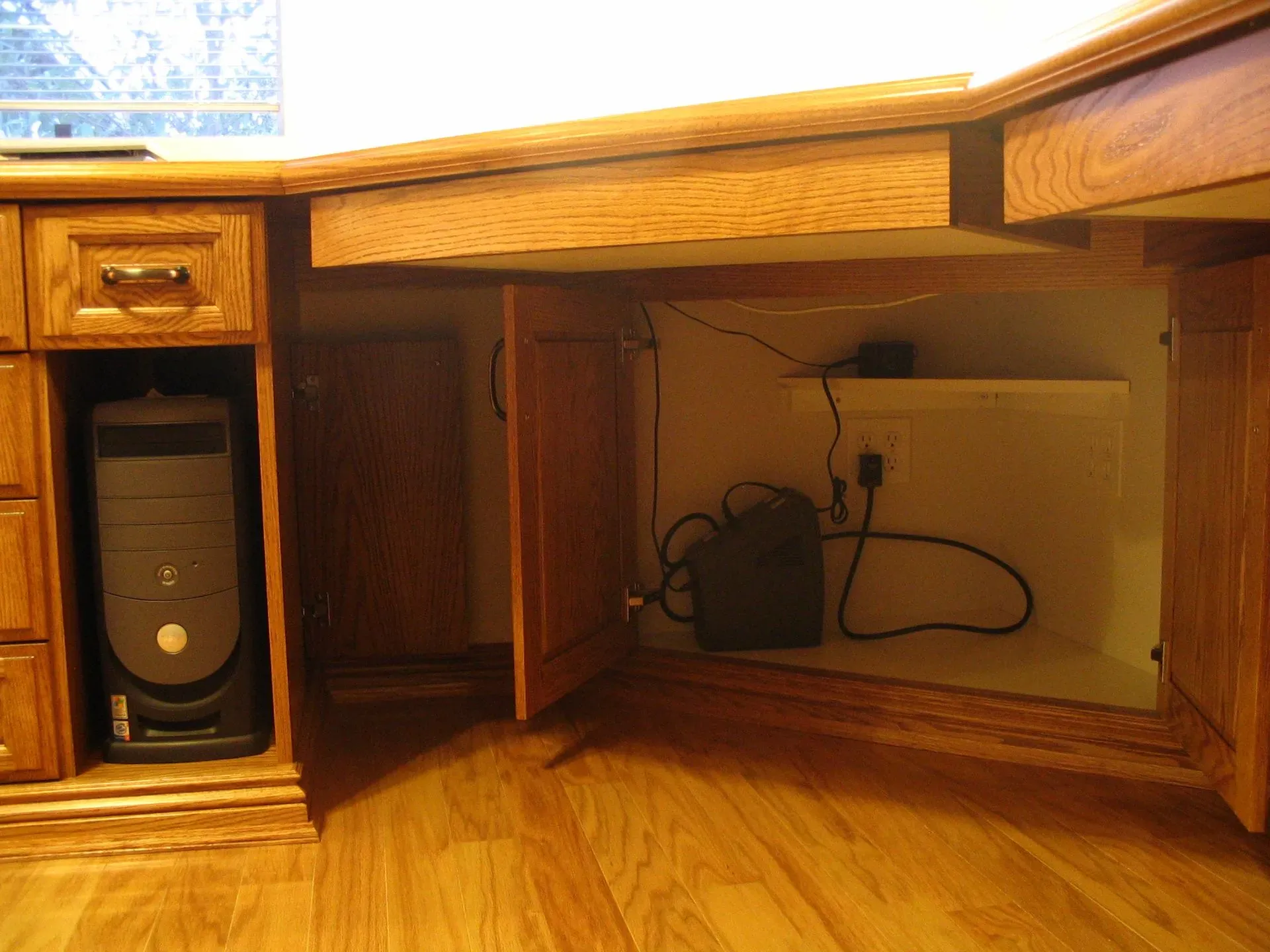 Wooden desk with open cabinets, showing a desktop computer tower and cables underneath.