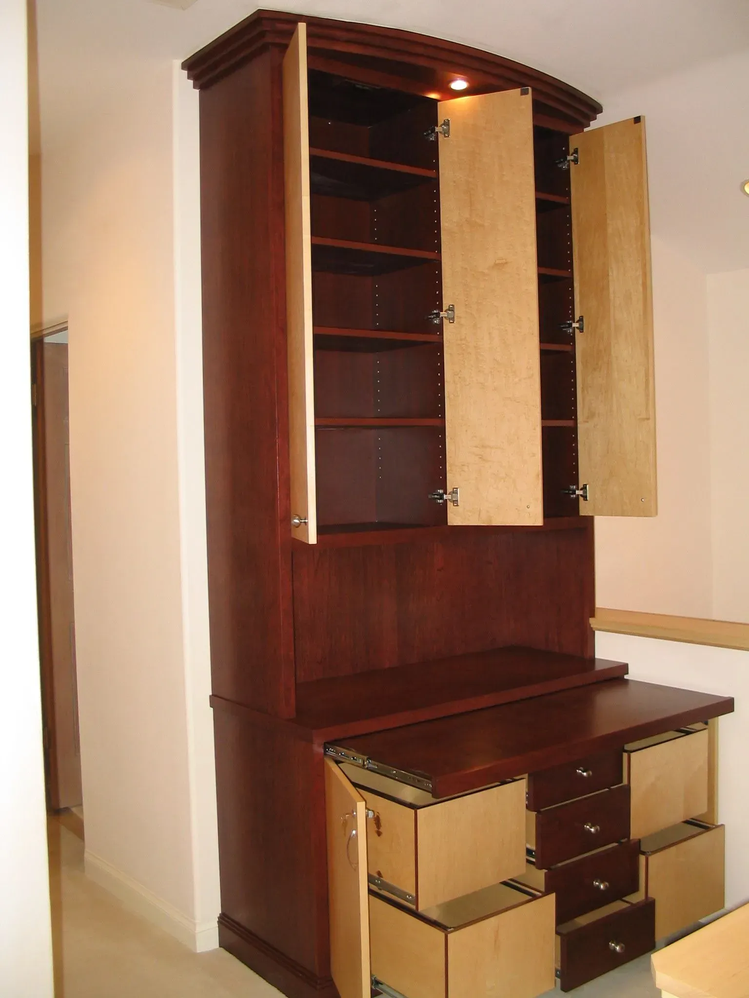 Wooden cabinet with open doors and drawers, showing a built-in desk and shelving inside a room.