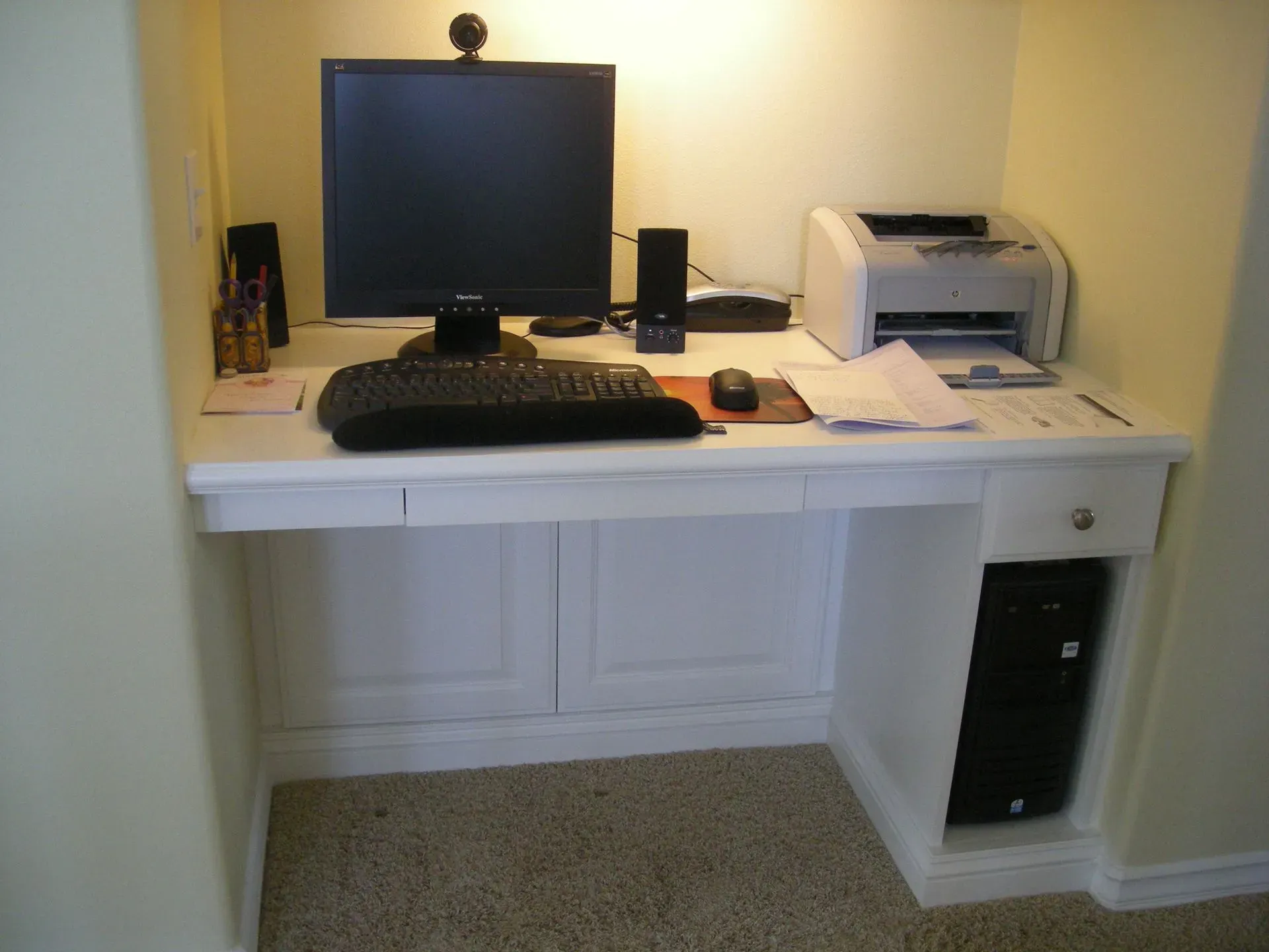Home office desk with computer monitor, keyboard, printer, and speaker against a wall.