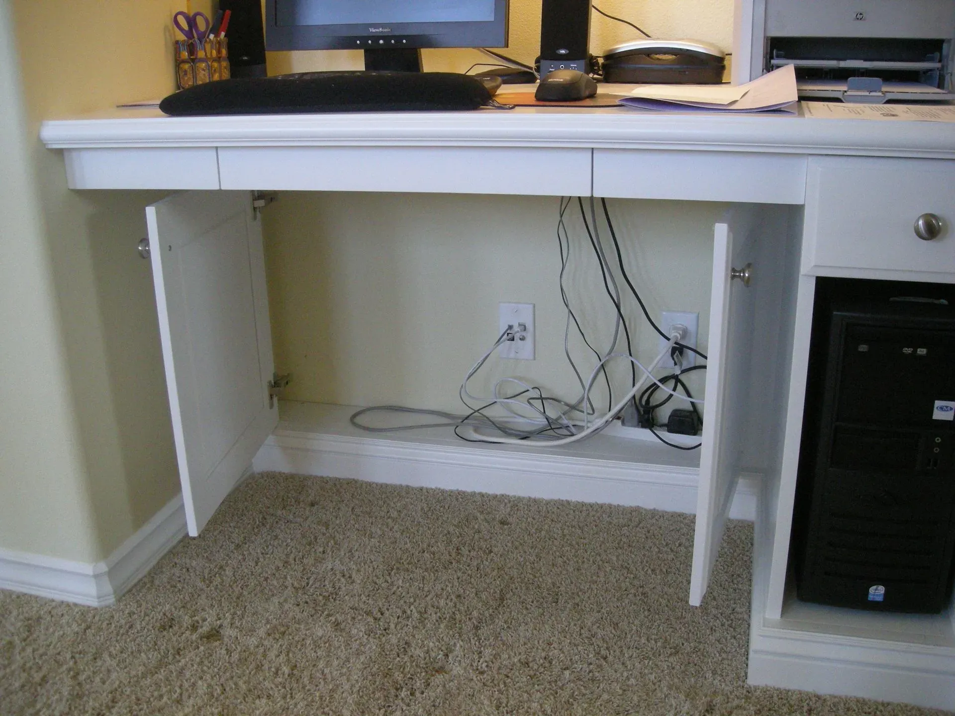White desk with shelves and tangled cables under it beside a black computer tower on carpet.