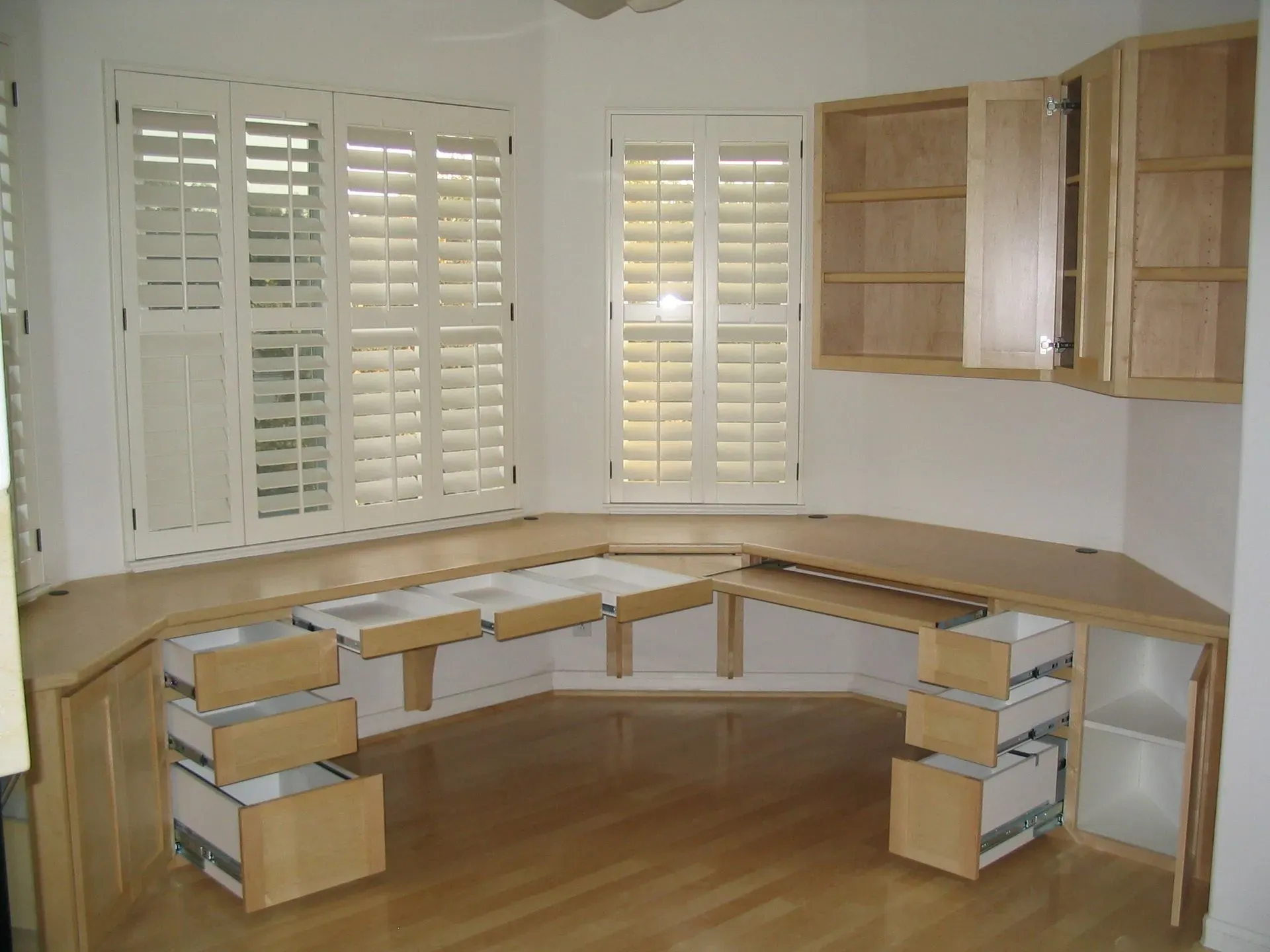 Curved wooden home office desk with drawers and upper cabinets in a bright room with white shutters