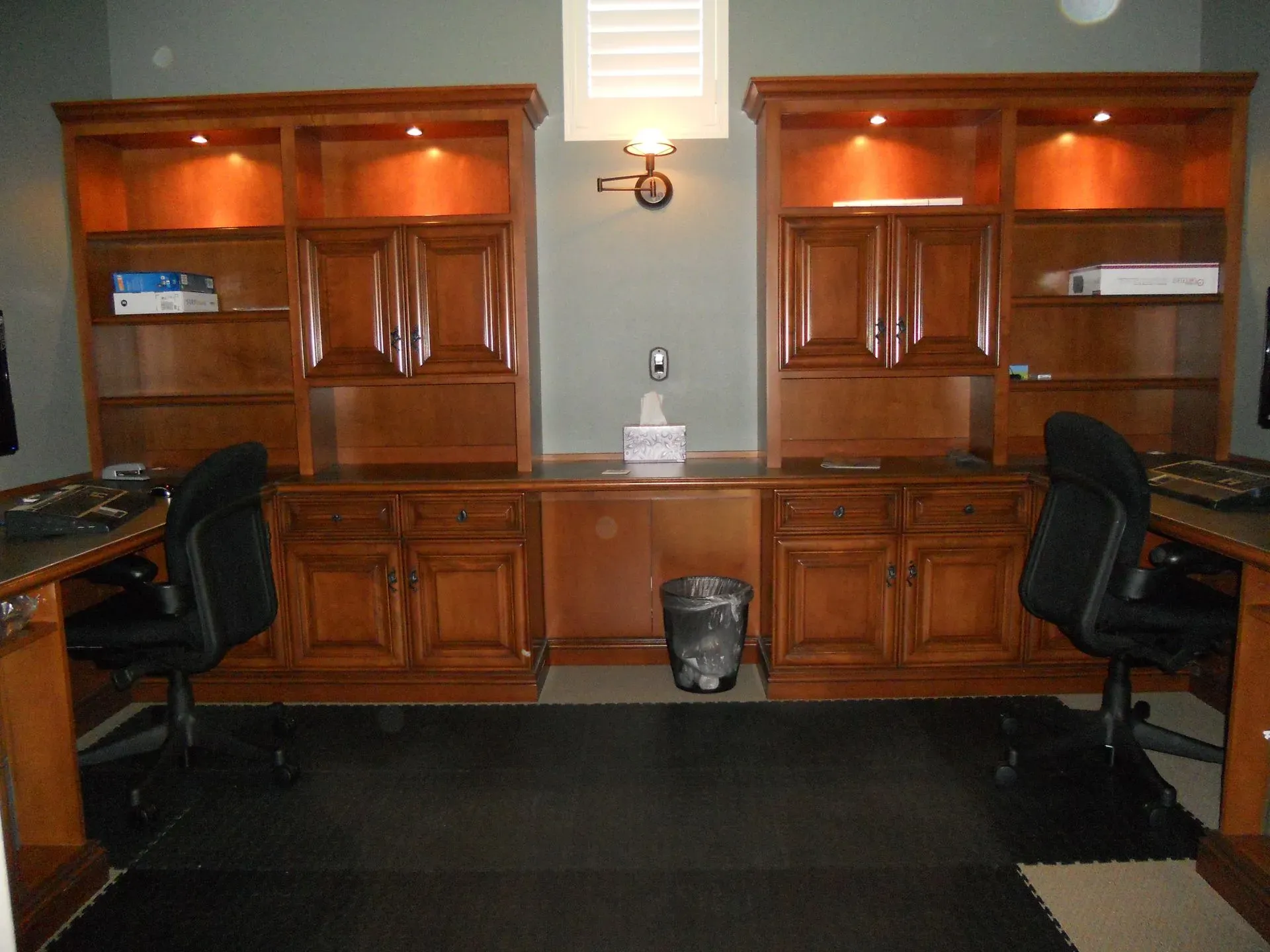 Wooden built-in desks with black chairs in a home office, lit by warm accent lights.