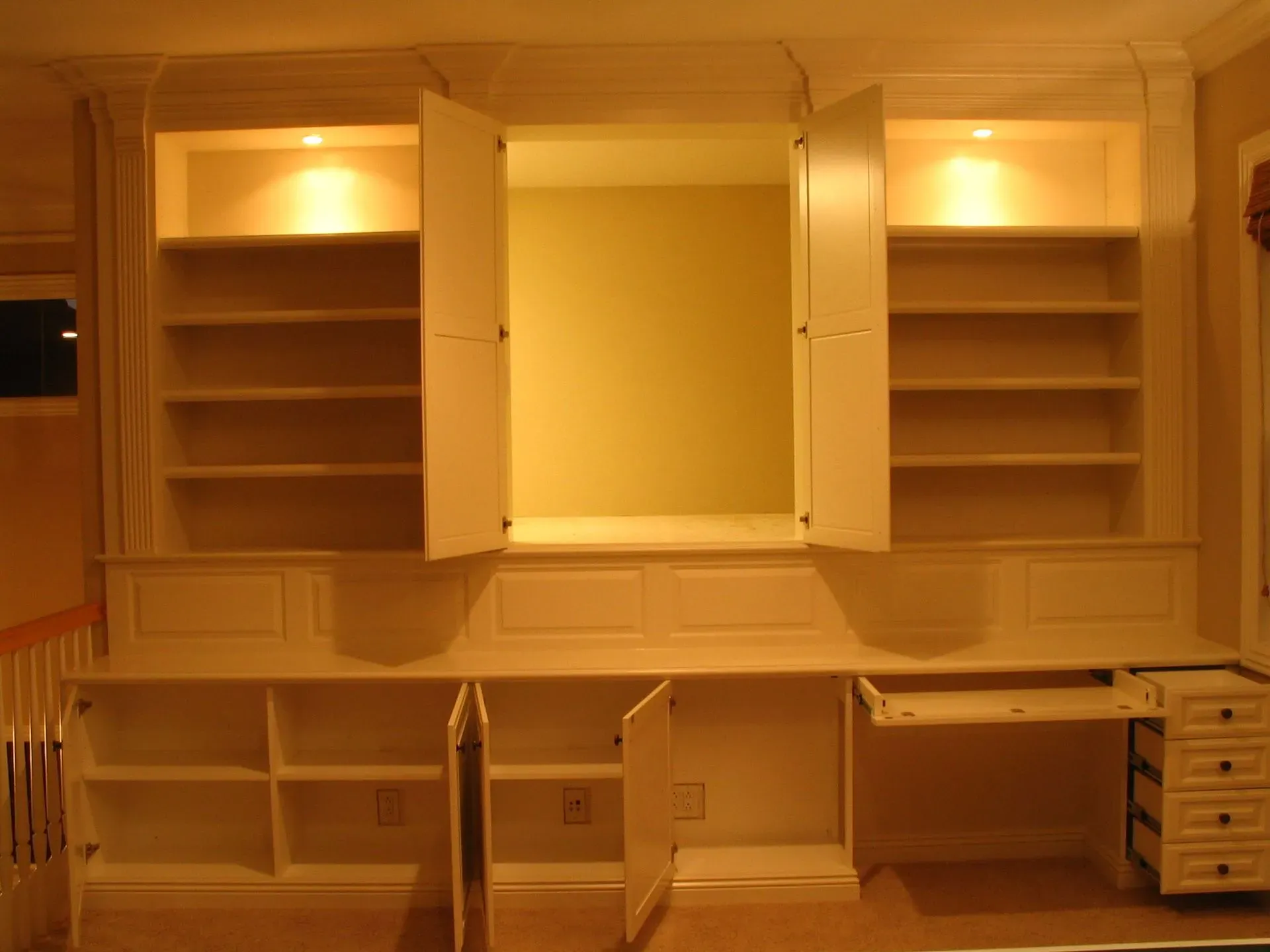 Built-in wall unit with open shelves, cabinets, desk, and warm overhead lighting in a room.