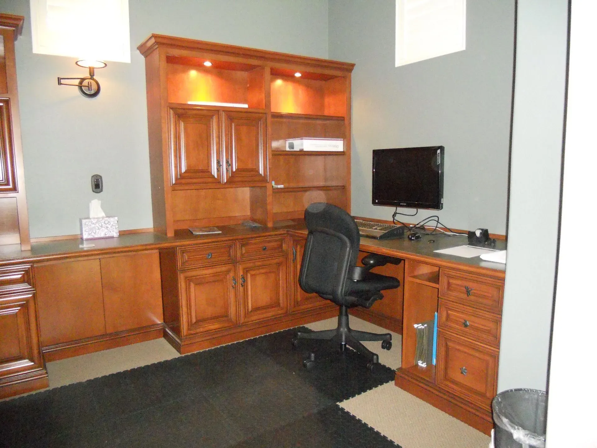 Home office with wooden desk and cabinets, computer monitor, black chair, and dark rug