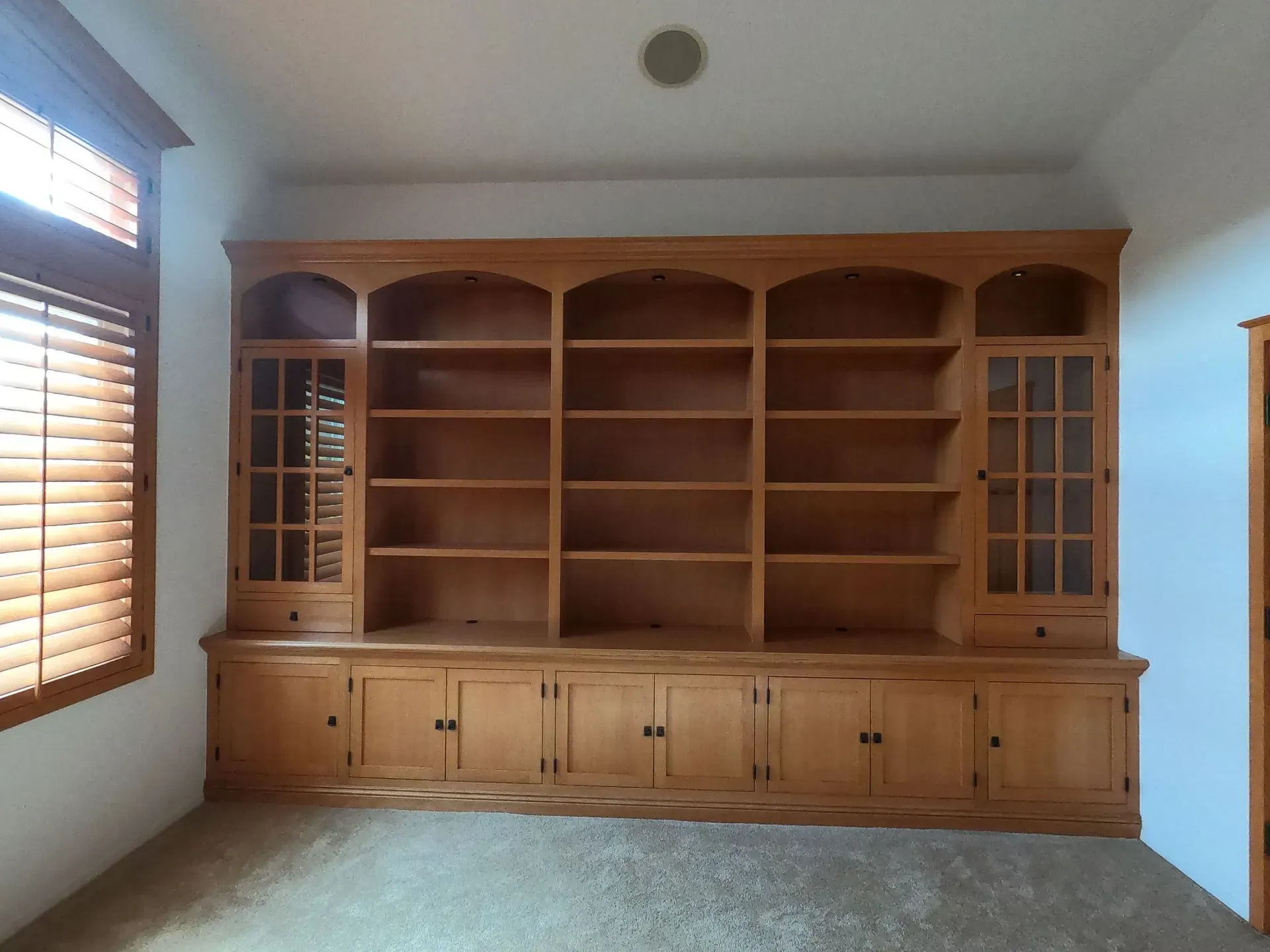 Built-in wooden wall bookcase and cabinet with shelves, in a bright room with windows.