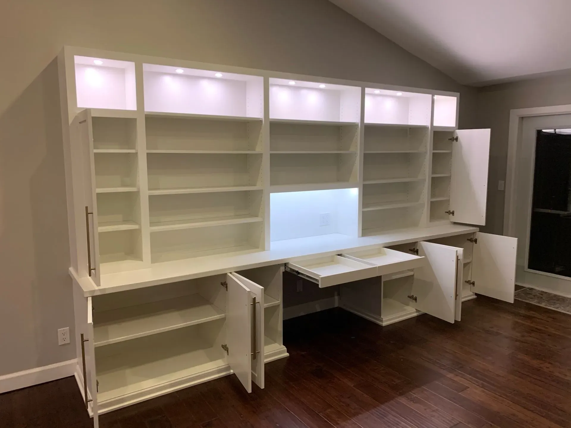 White built-in office wall cabinets and desks with shelving, lit display panels, in a room with wood floors.