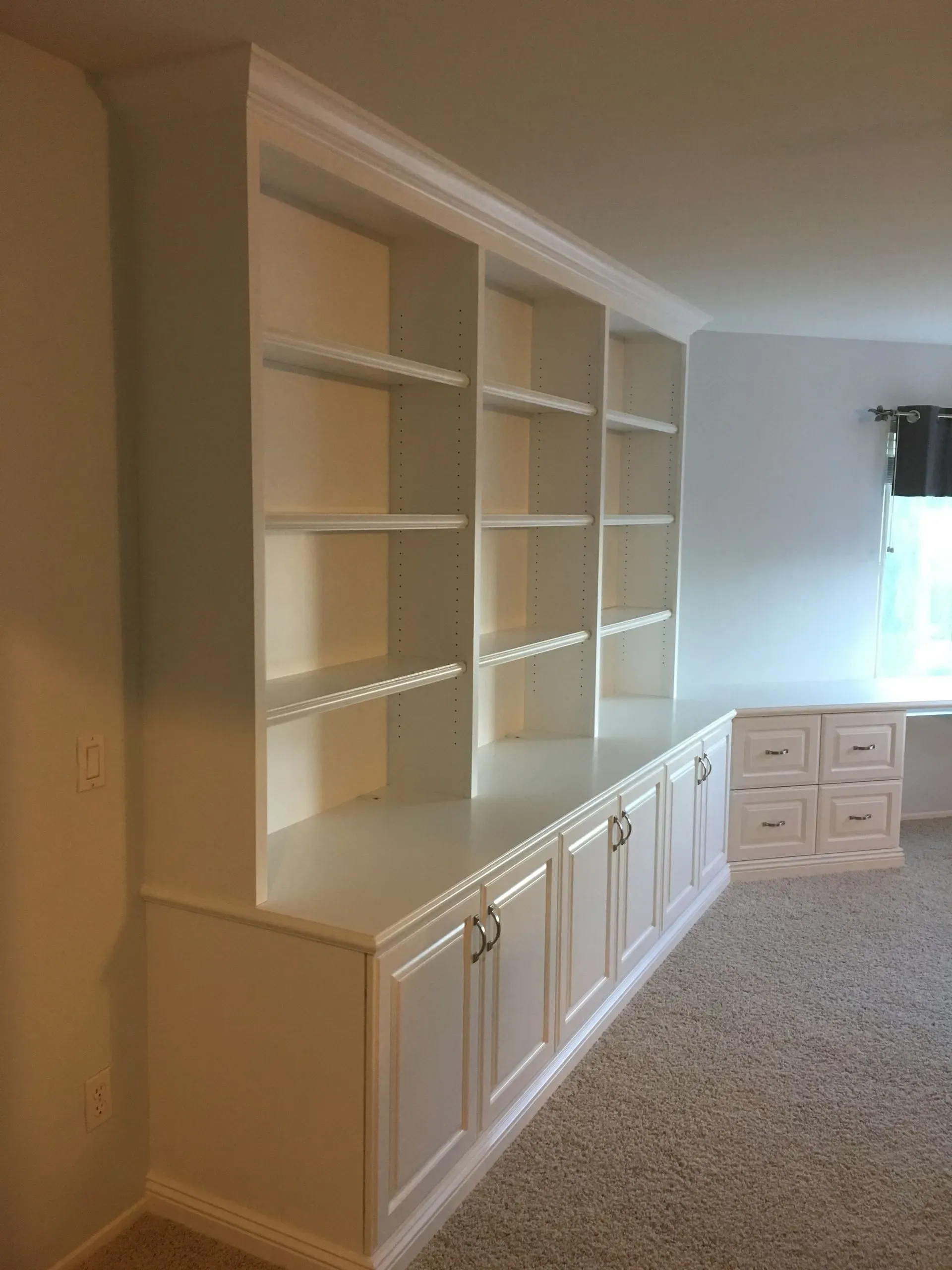 Built-in white shelves and cabinets along a beige-carpeted wall in a bright room