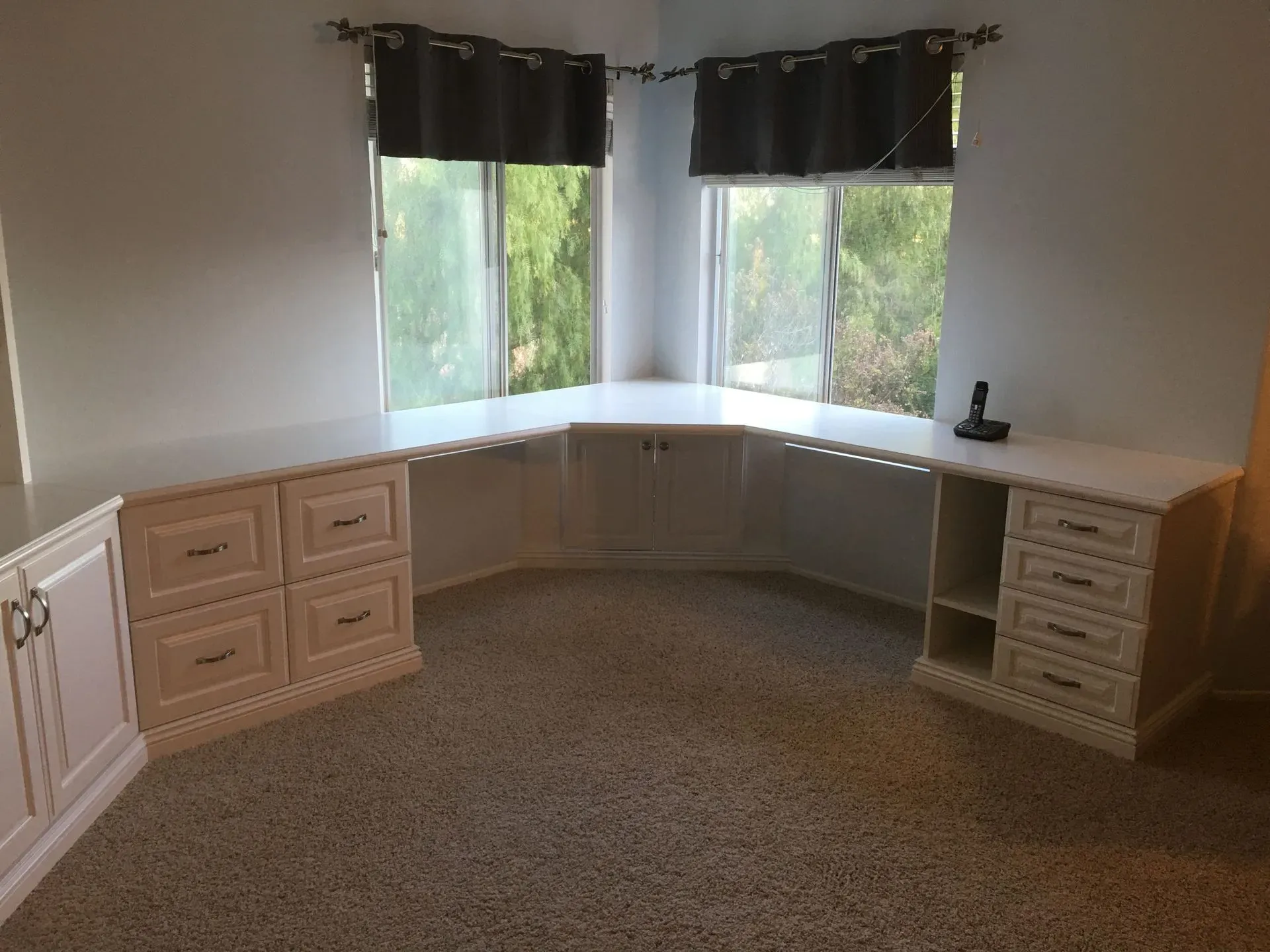 Empty carpeted office with built-in corner desk, light wood cabinets, and windows with dark curtains.