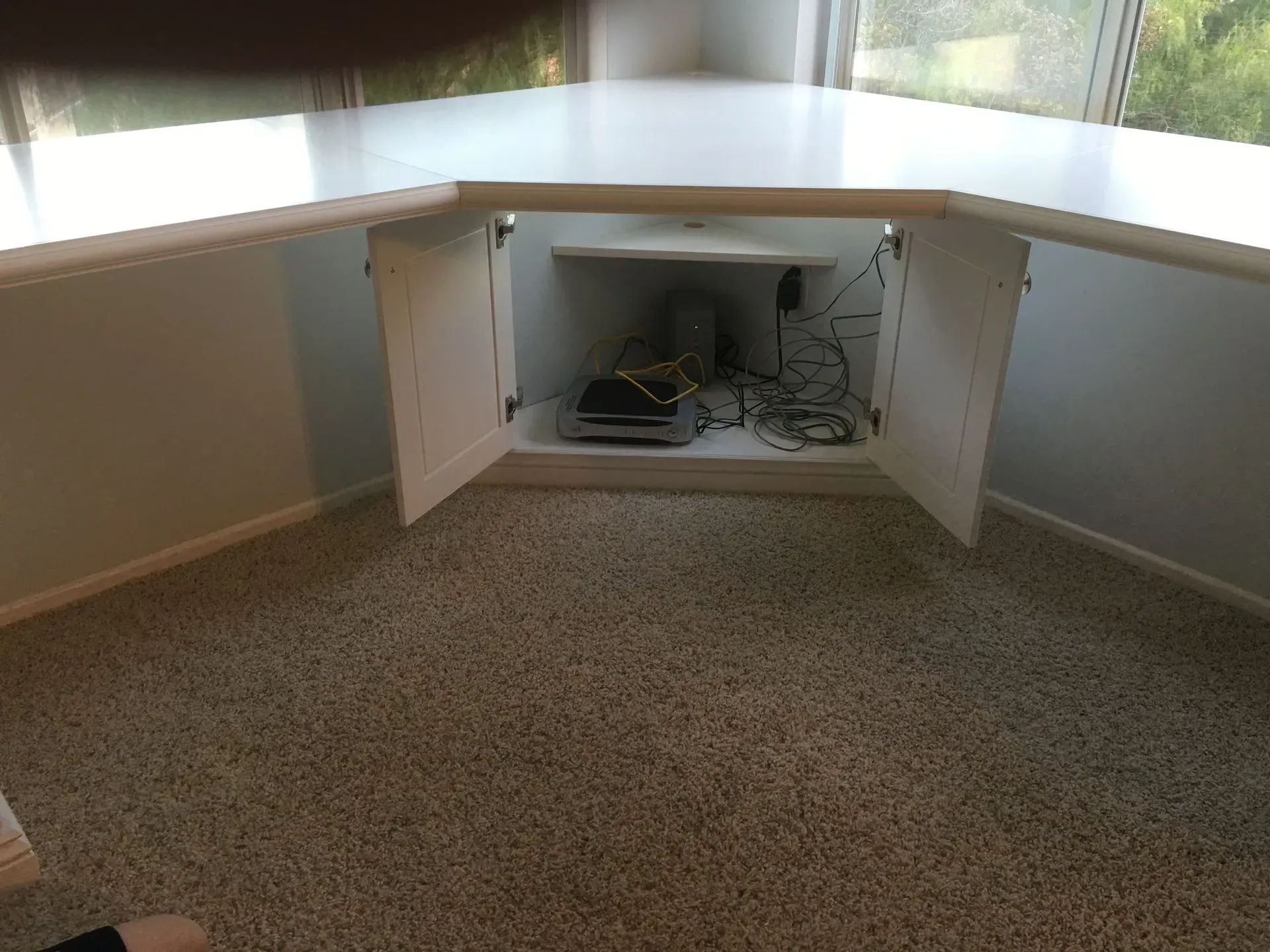 Corner office nook with open cabinet under a white countertop and visible cables, beside carpet and windows.