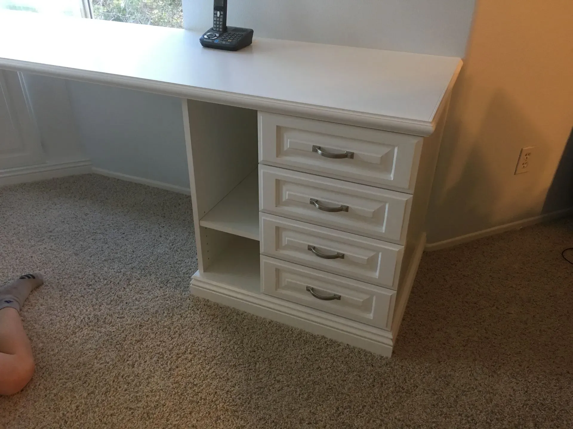 White desk with four drawers and open shelf in a carpeted room, with a lamp on top.
