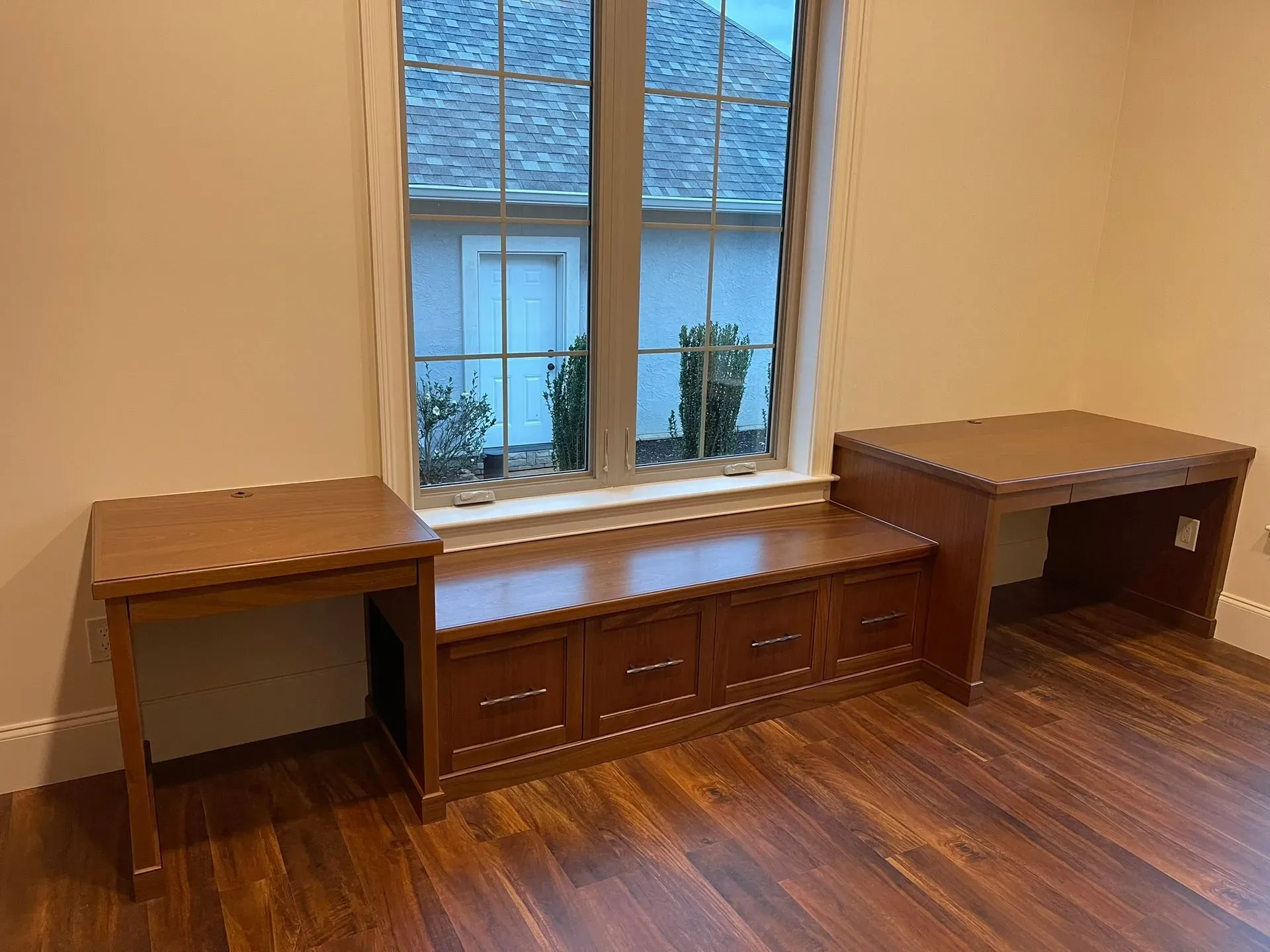 Wooden furniture arranged beneath a window in a beige room, with a low cabinet centered between two desks.