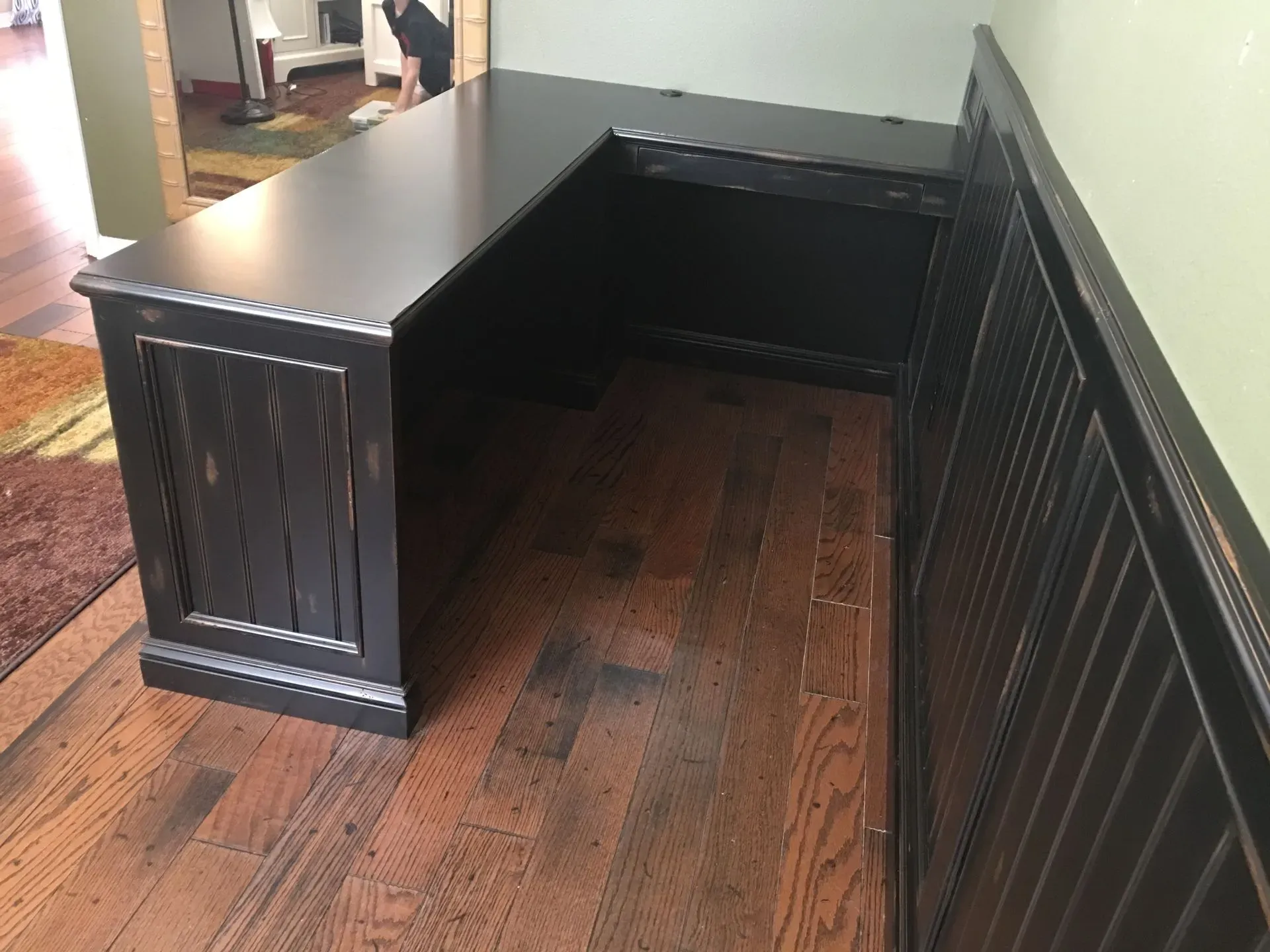 Black L-shaped desk with a single pedestal in a hardwood-floored room, viewed from the corner.