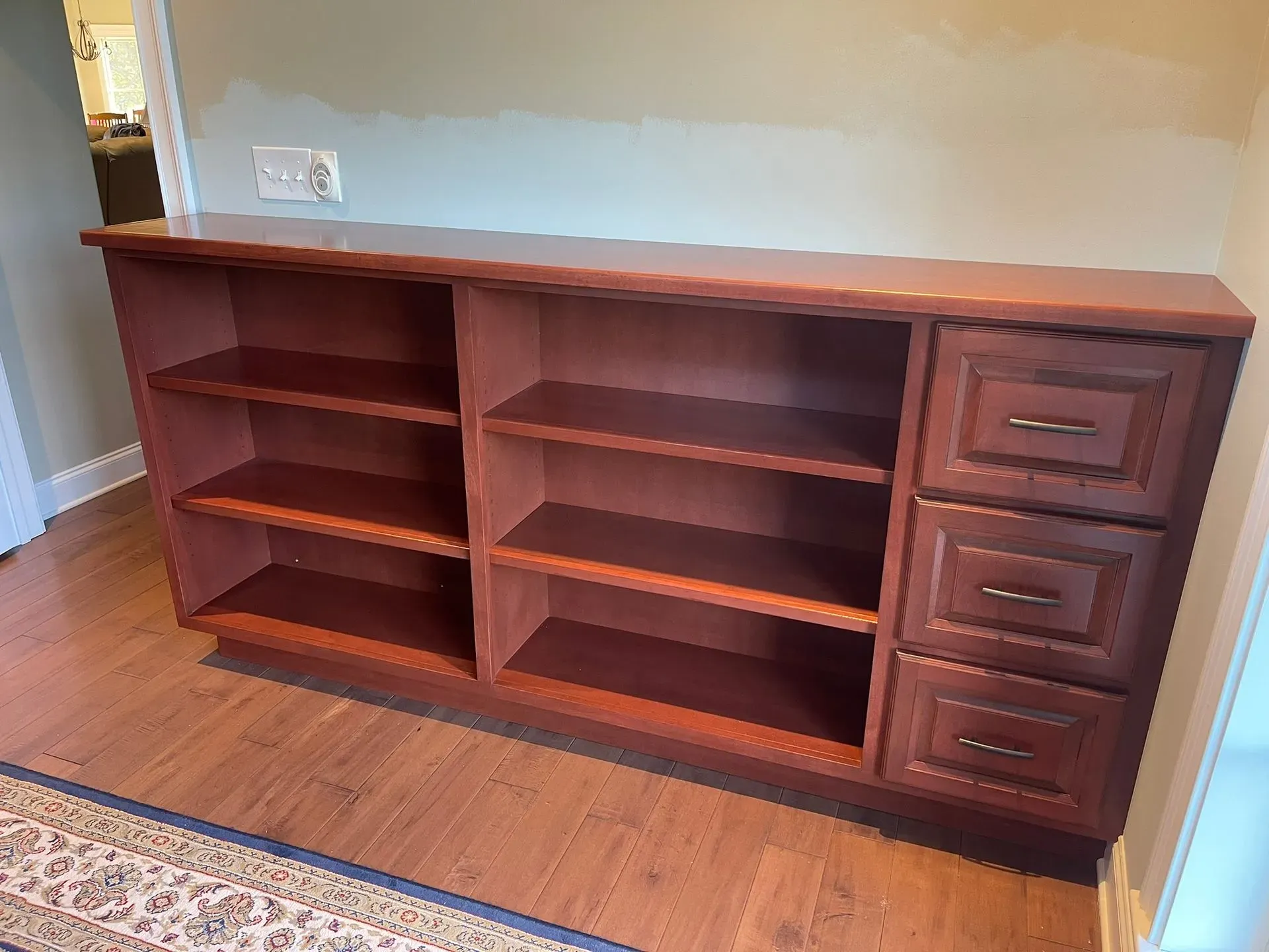 Cherry wood bookshelf with open shelves and three drawers against a beige wall.