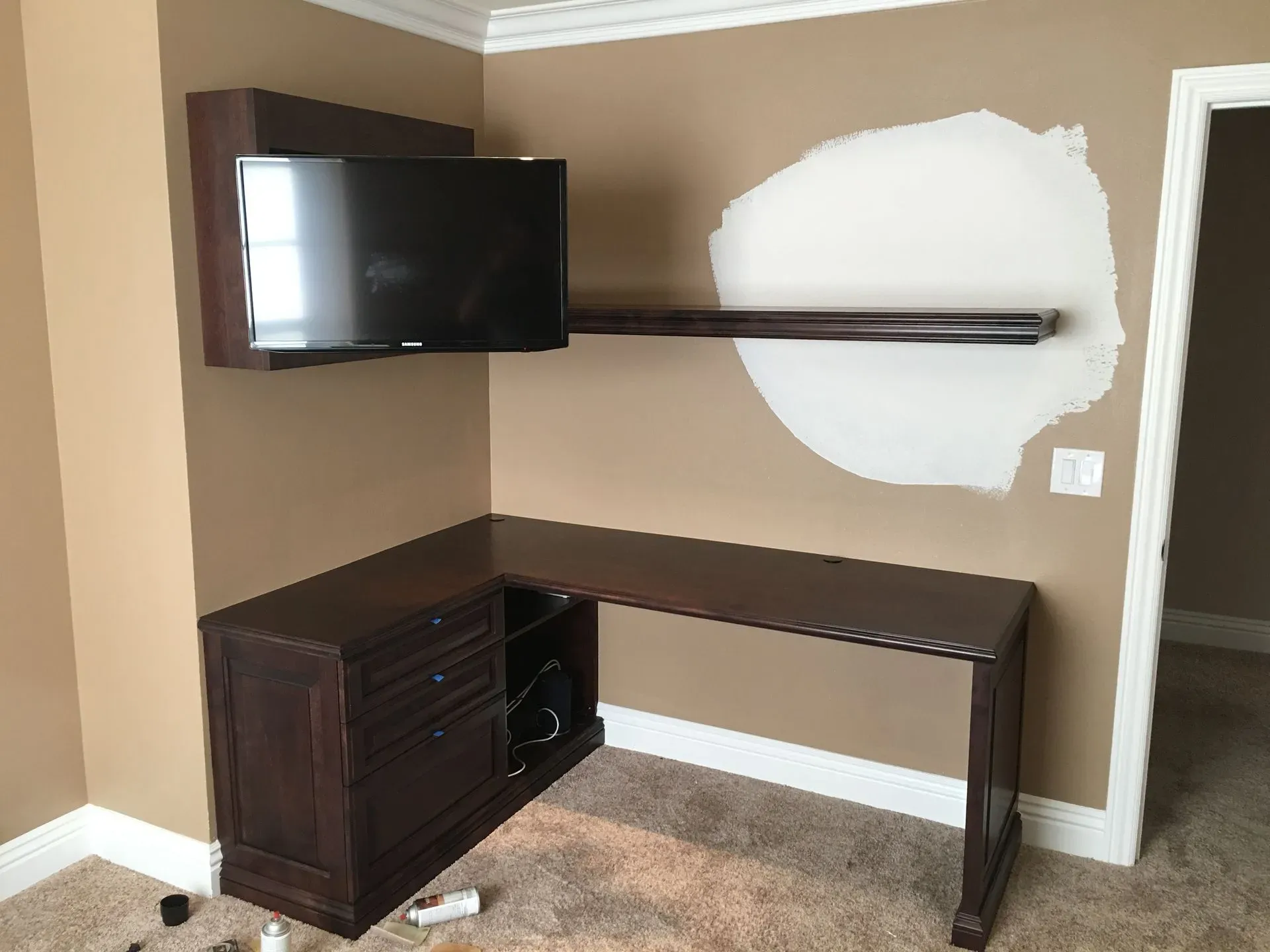 Corner wall-mounted TV above desk with dark wood cabinets and white wall patch in a beige room