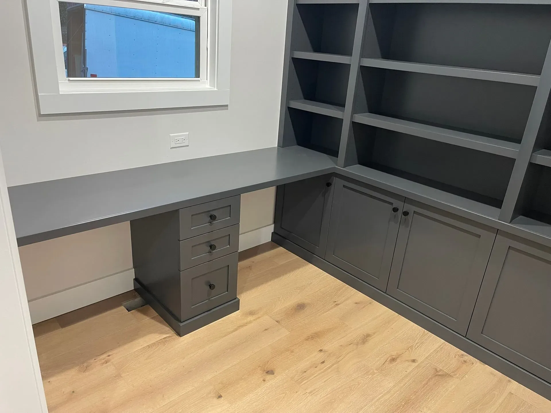 Built-in gray desk and shelves in a small office nook with wood floor and window