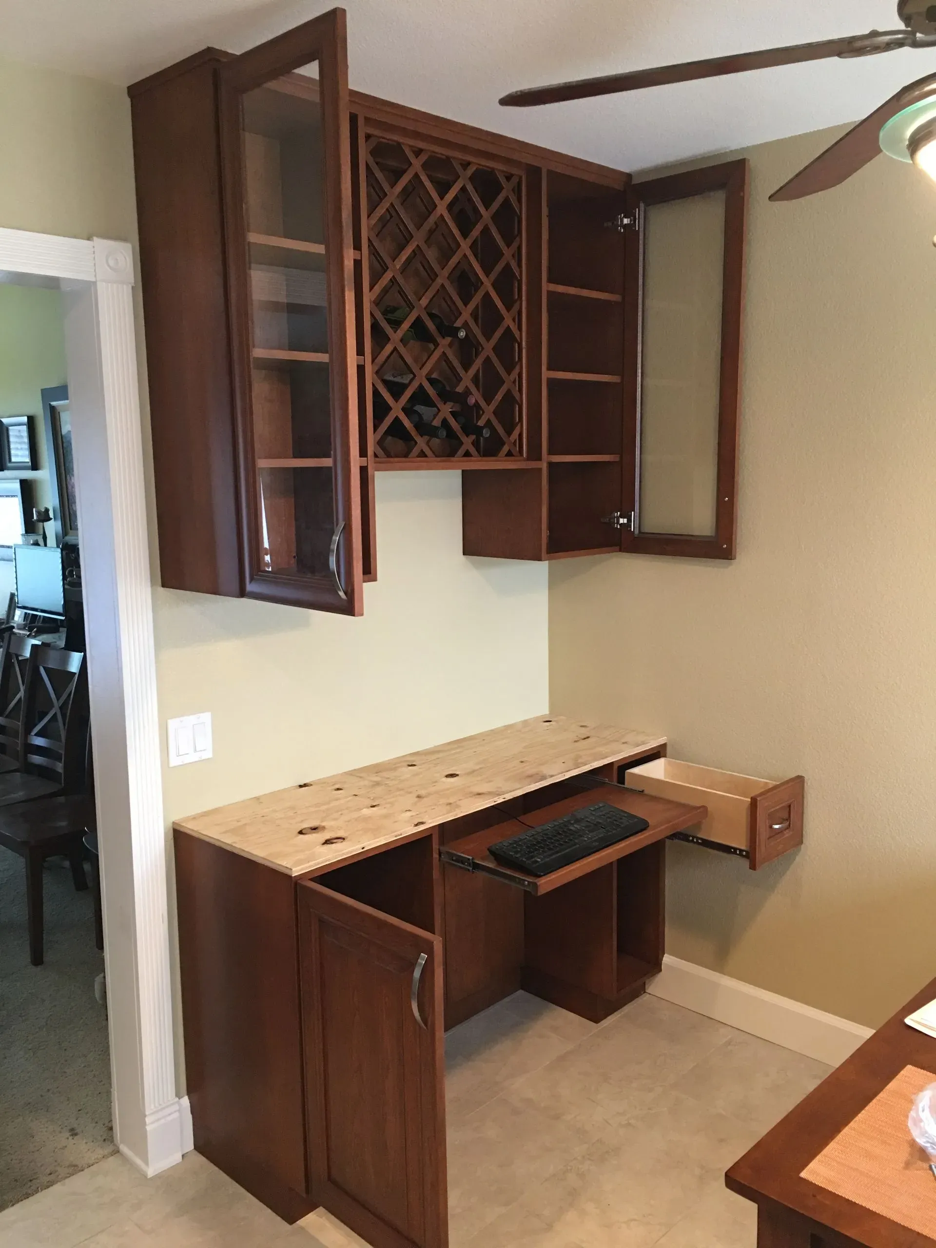 Wood desk with upper cabinets and wine rack in a beige home office corner, cabinet doors open.