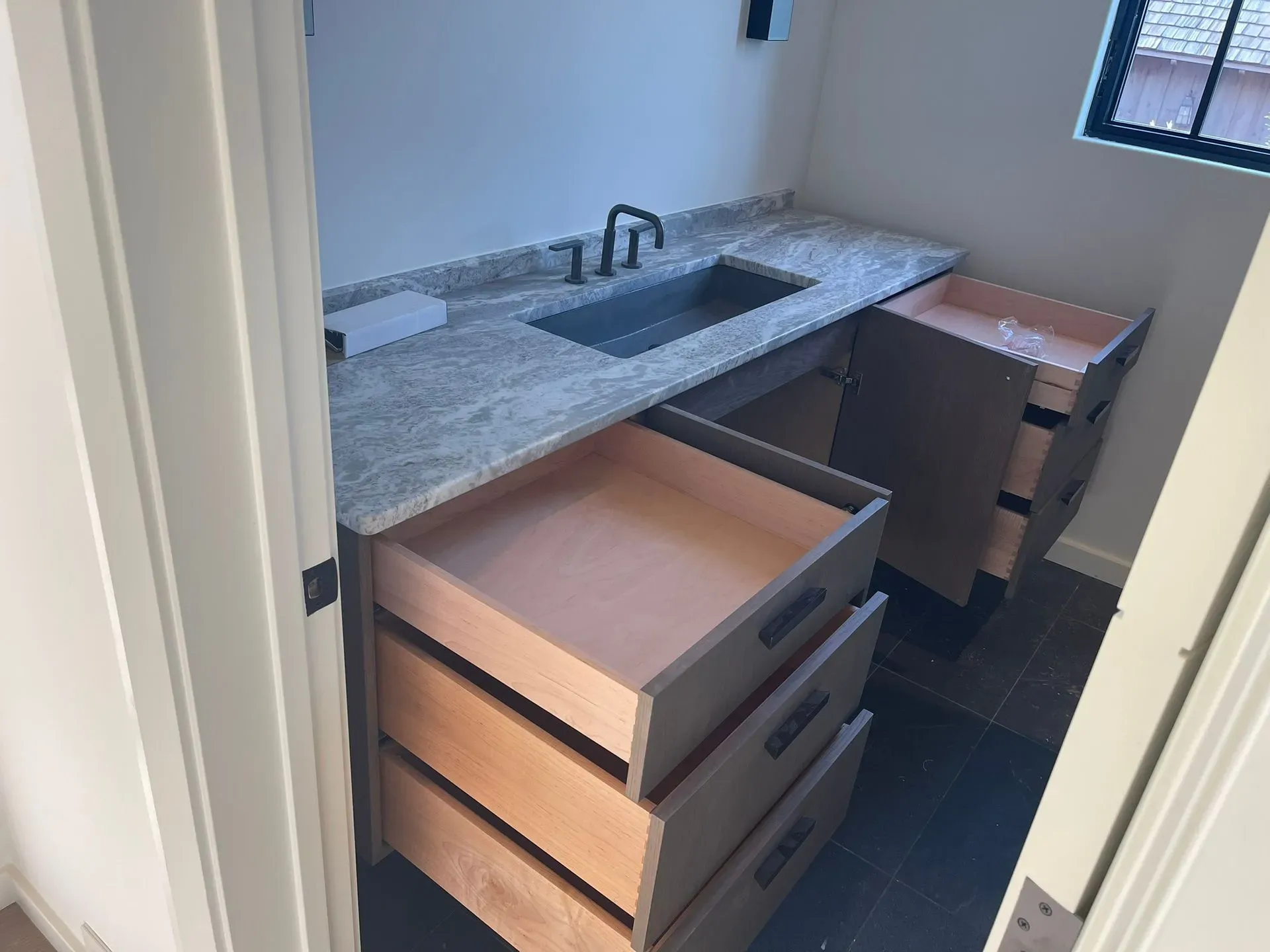 Small bathroom vanity with granite sink, open drawers, and a window above the counter.