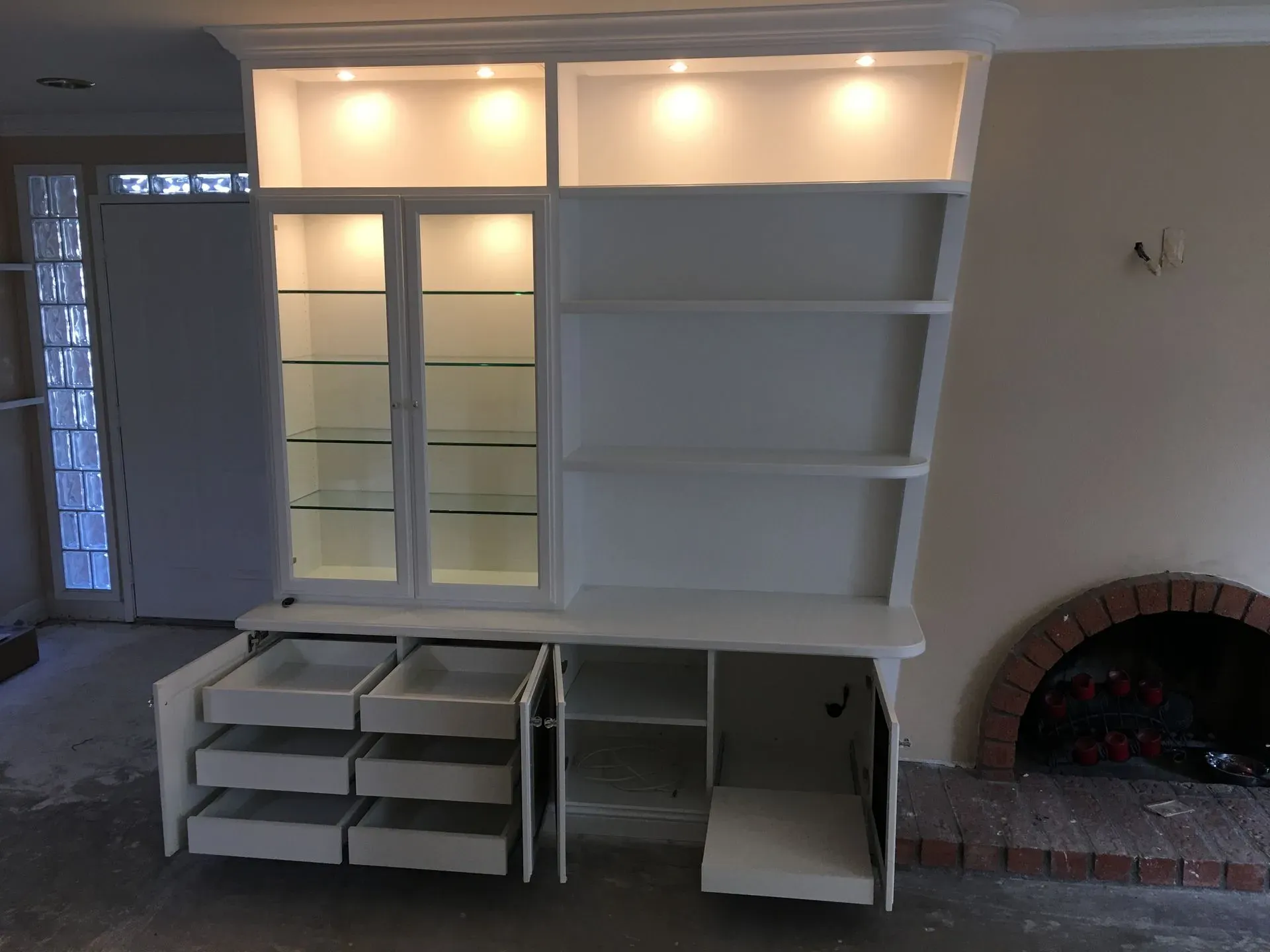 Built-in white shelving unit with lit cabinets, drawers, and an arched brick fireplace nearby
