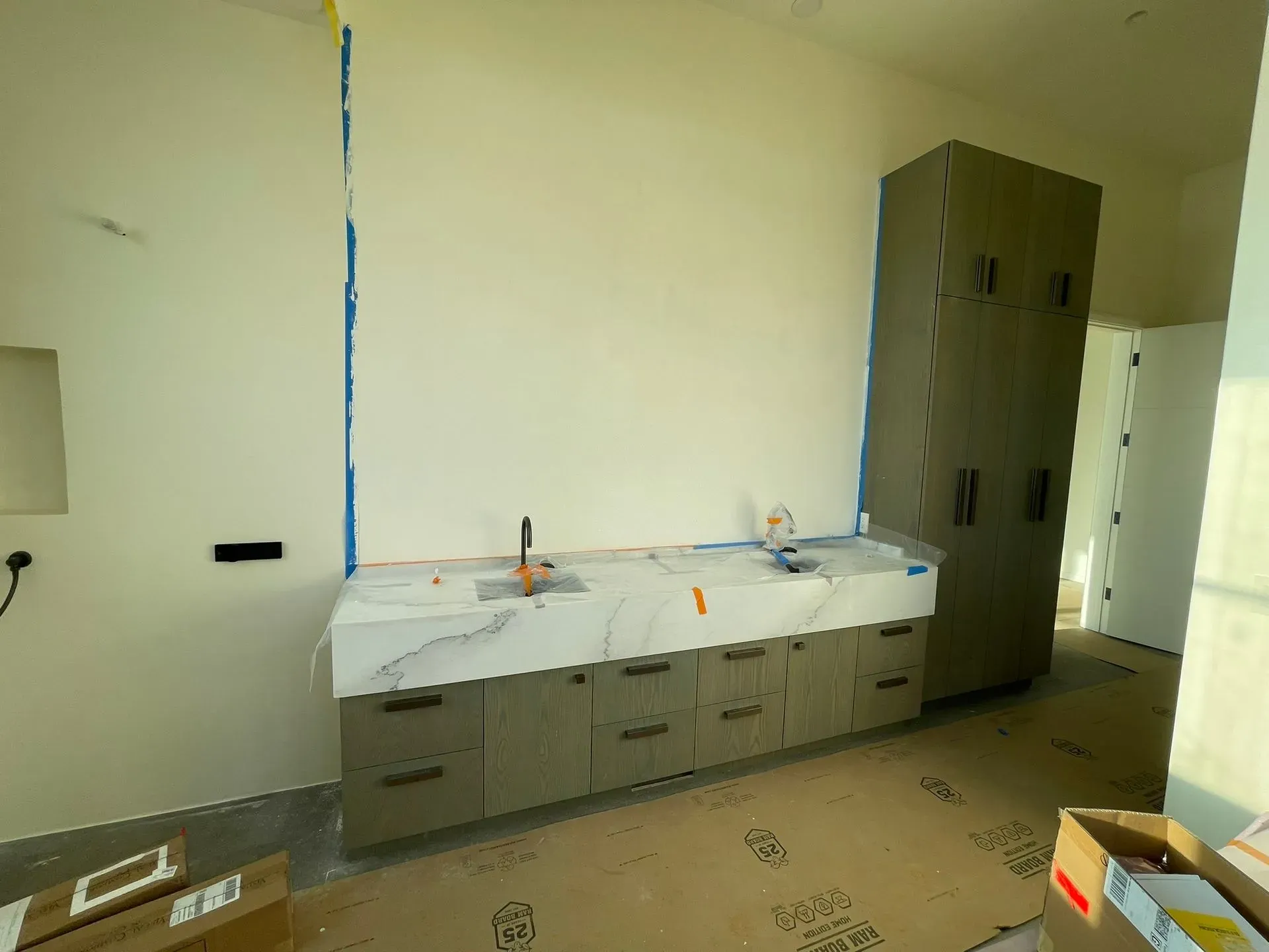 Kitchen under renovation with cabinets, covered countertop, and a tall storage cabinet against a bare wall.
