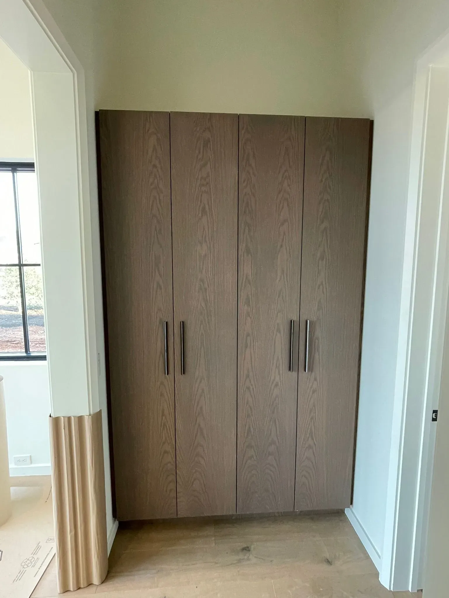 Built-in brown wooden wardrobe in a bright hallway with white walls and a window.