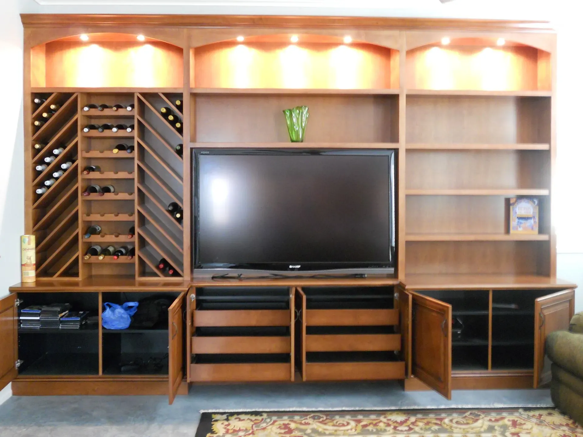 Wood entertainment center with TV, wine rack, cabinets, and shelves under warm spotlights.